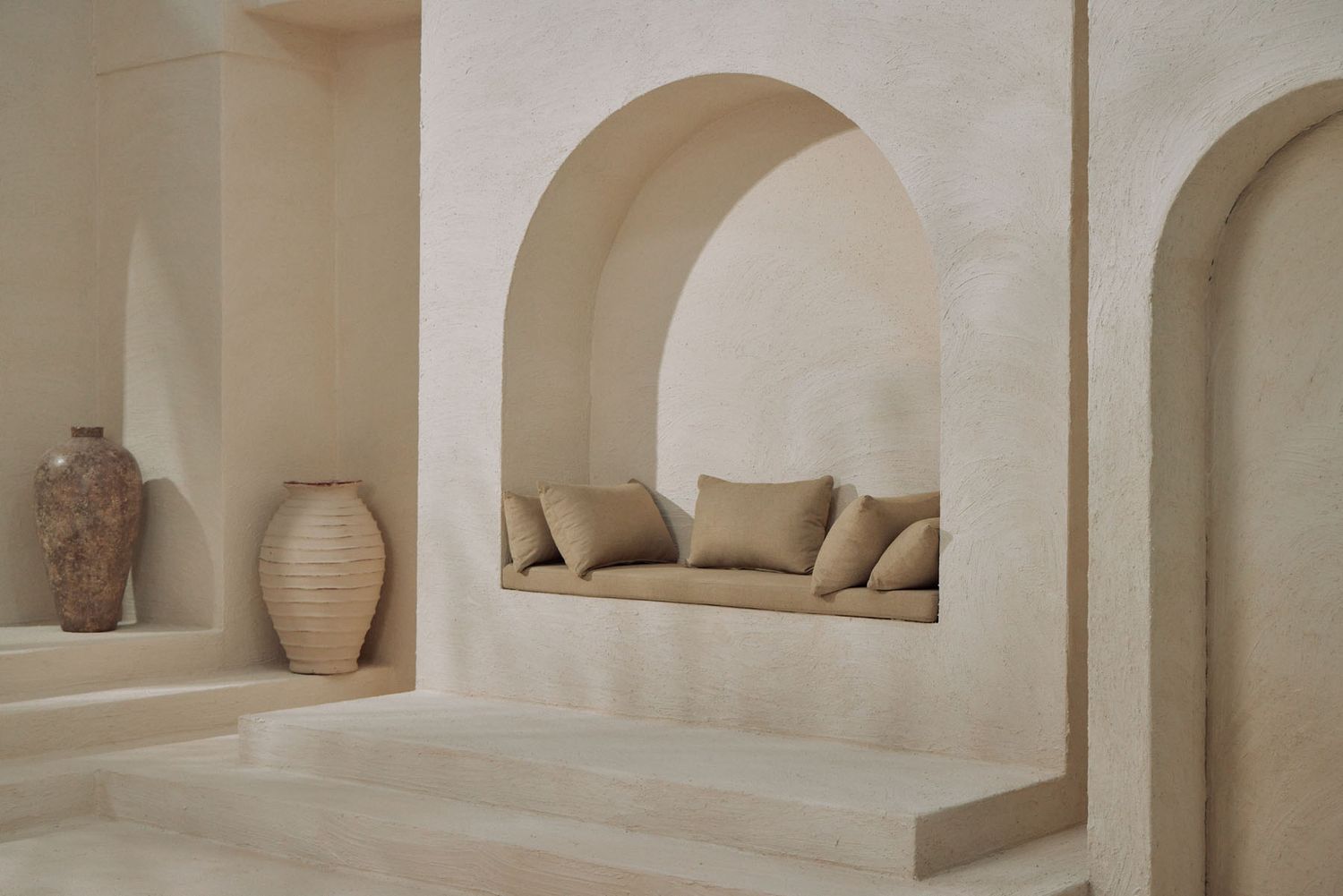 Minimalist beige plaster interior with arched niche seating, neutral linen cushions, and decorative terracotta vase.
