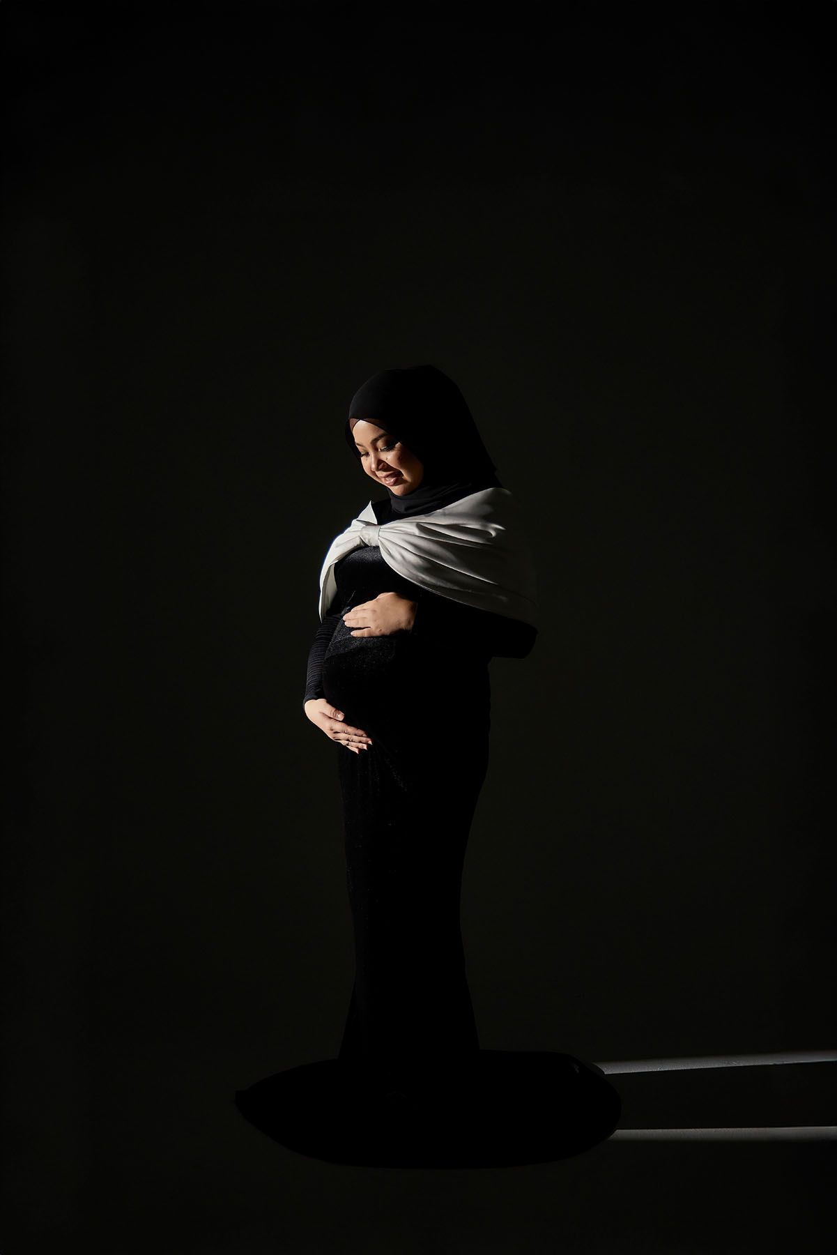 Pregnant woman in black dress and white hijab cradling her baby bump in a dramatic dark studio portrait.