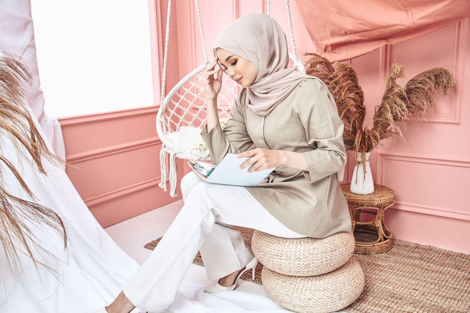 Woman in beige hijab and outfit writing on clipboard while sitting in pink decorated room.