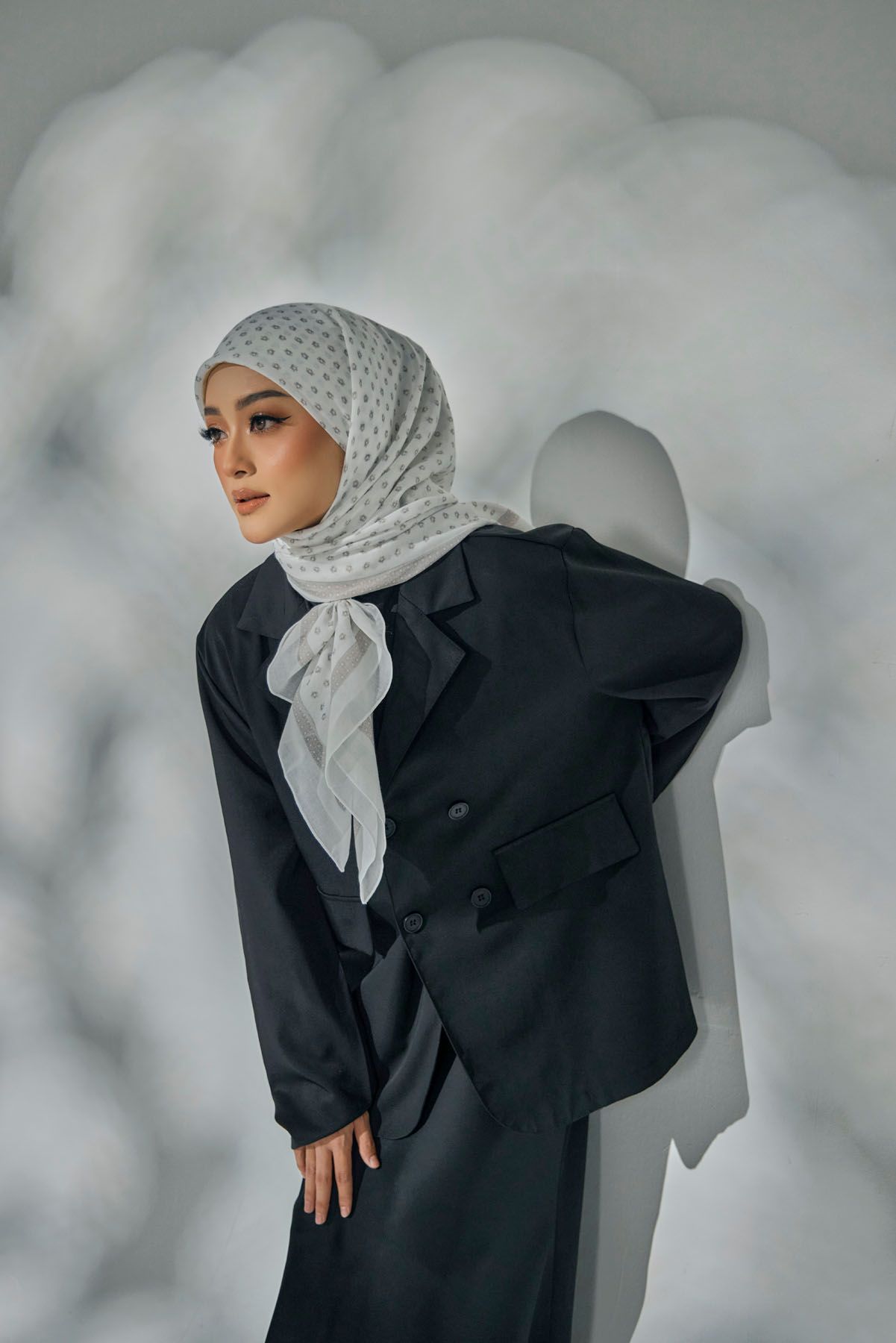 Fashionable Muslim woman in black blazer suit with a white polka dot hijab against a cloudy backdrop.