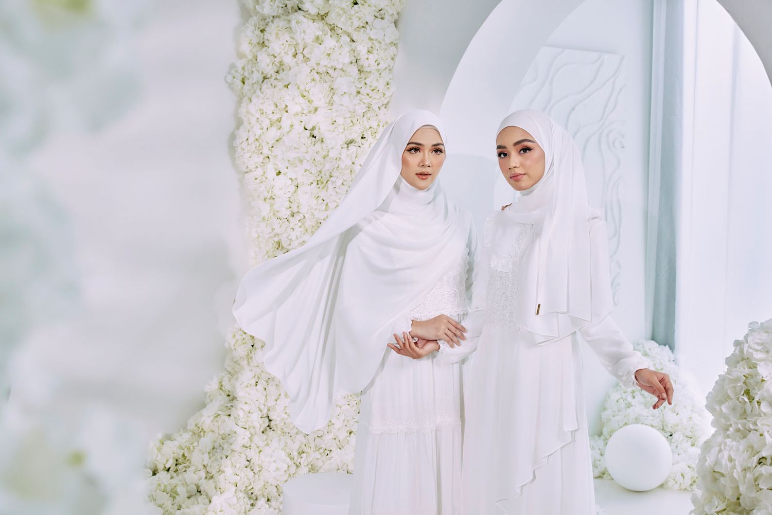Two elegant women in white hijabs and dresses posing in a white floral arch setting.