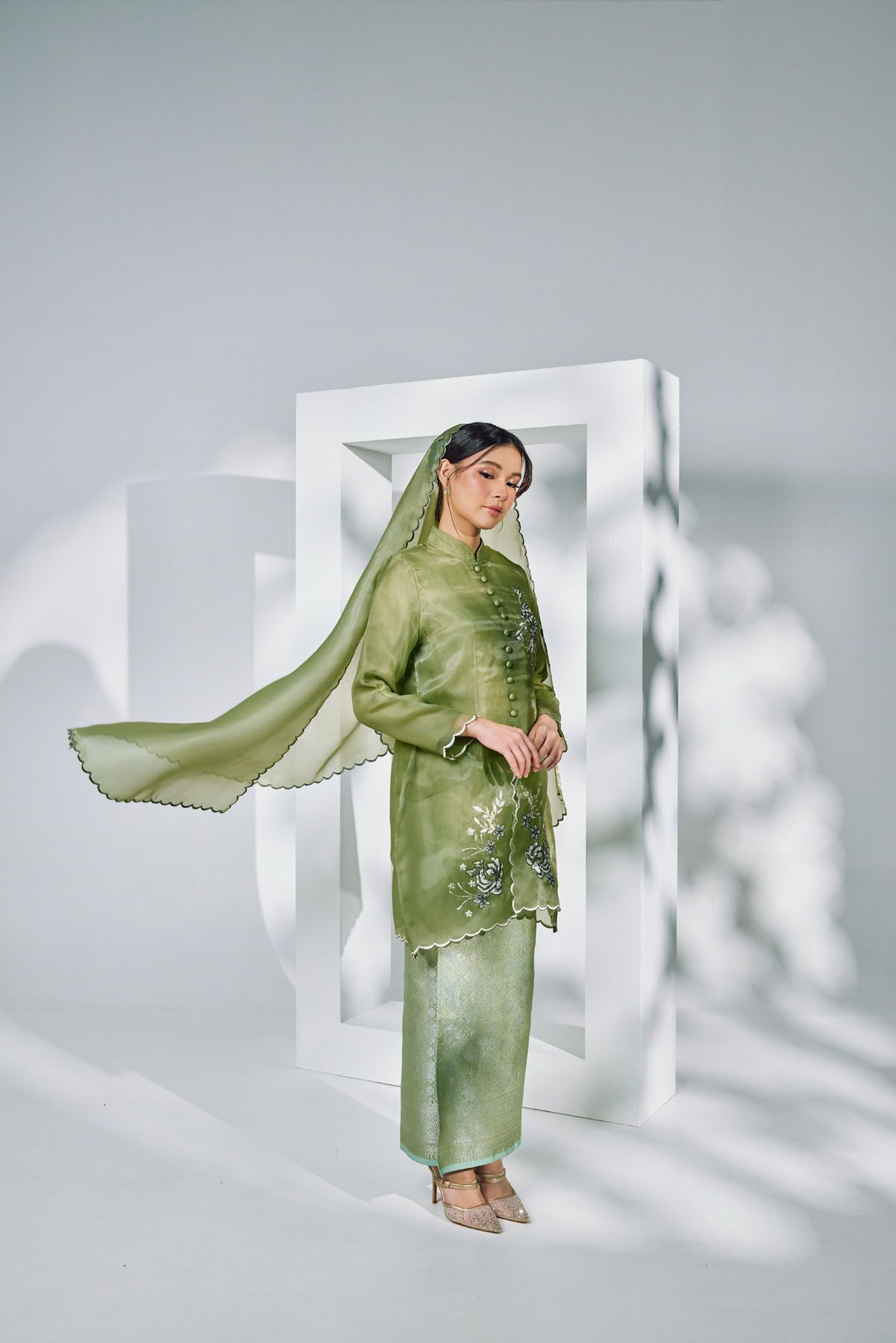 Woman in sage green embroidered kebaya with flowing dupatta posing near white geometric props.
