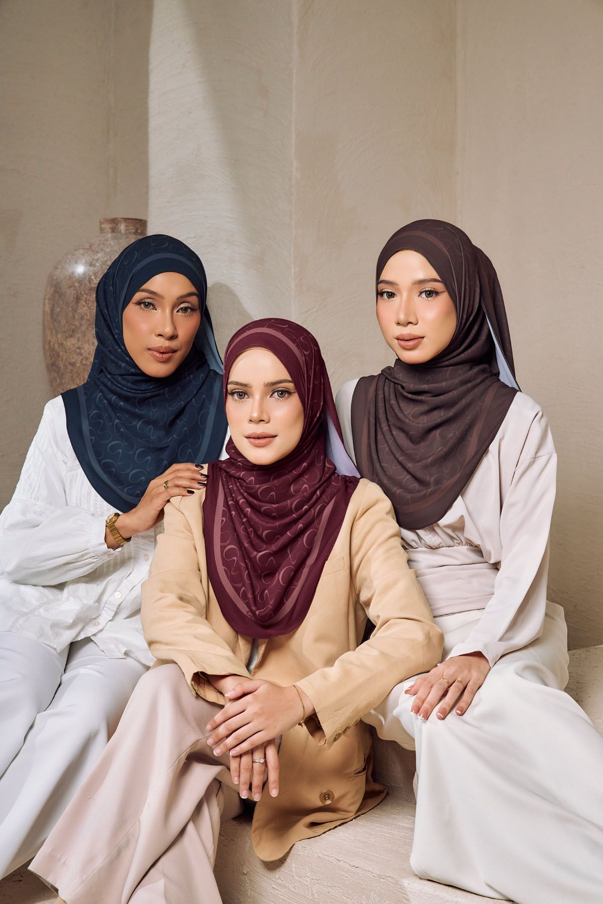 Three women in navy, burgundy, and brown hijabs pose together in elegant neutral setting.