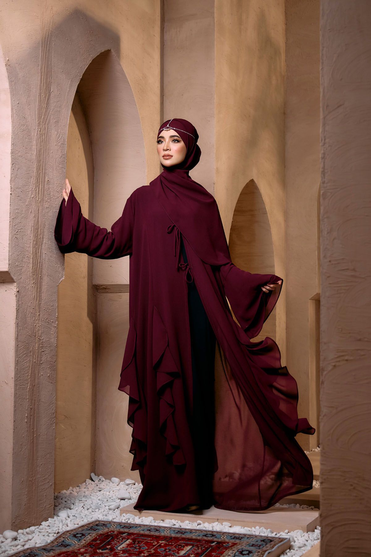 Woman in deep burgundy flowing abaya and hijab posing in ornate arched architectural setting.