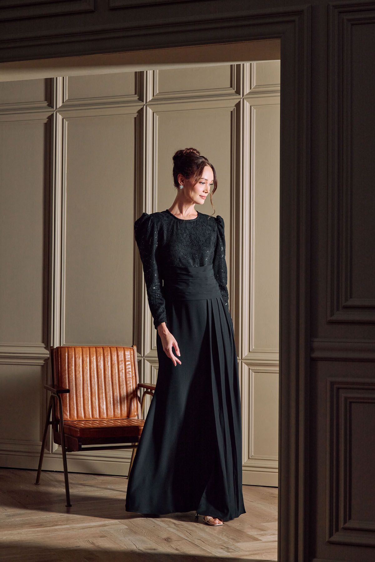 Woman in elegant black long-sleeve floor-length gown with lace bodice standing in a classic paneled room.