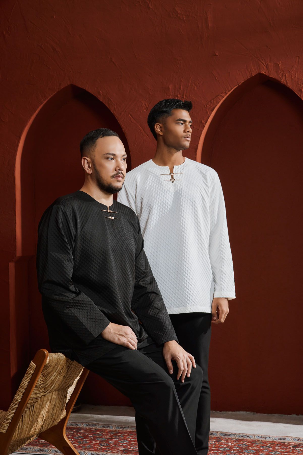 Two men in black and white quilted baju melayu posing against a red terracotta arched backdrop.