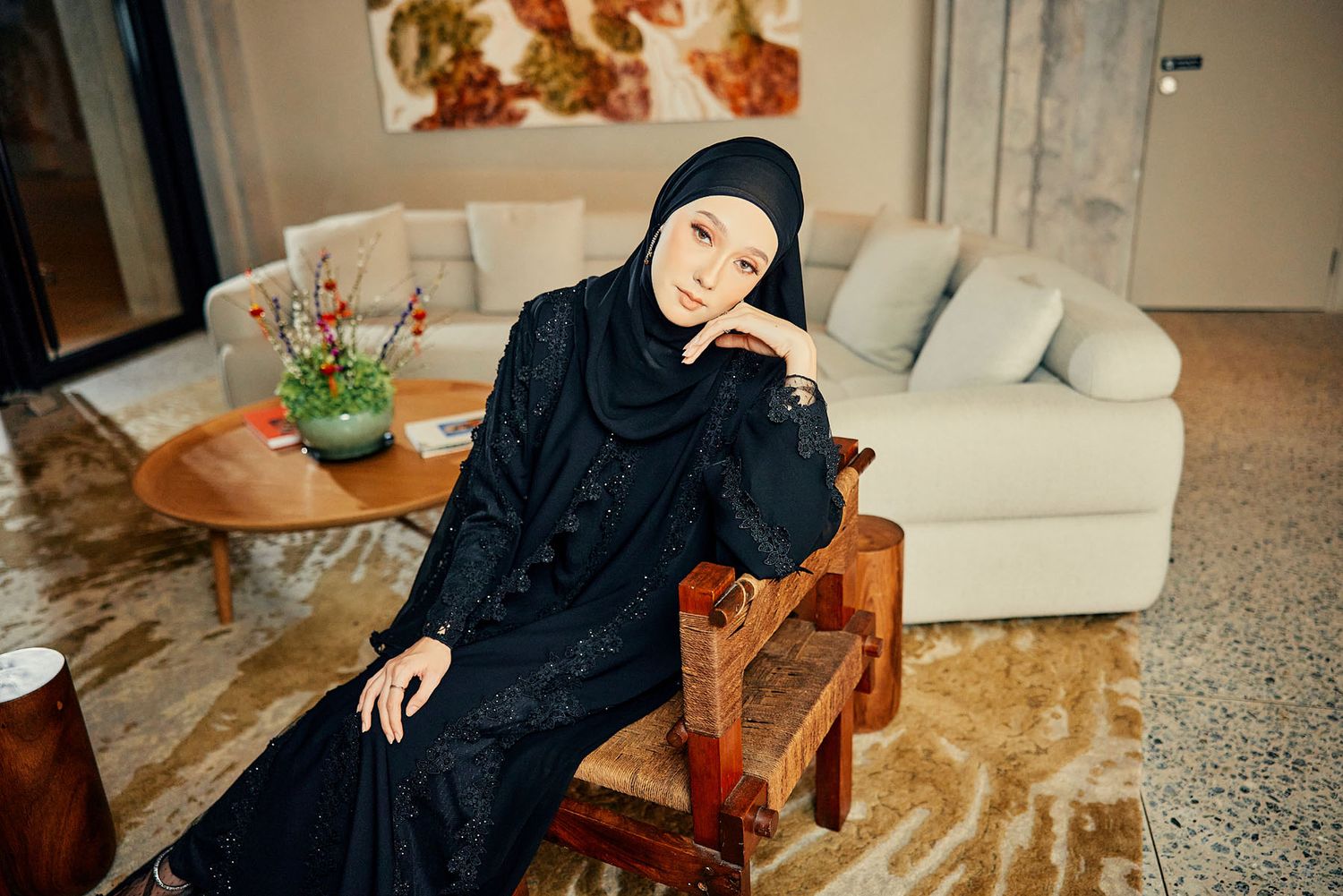 Woman wearing black sequined abaya and hijab seated on a wooden chair in a modern living room setting.