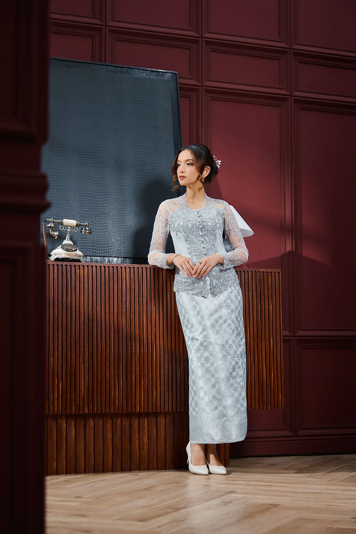 Woman in silver lace kebaya and skirt stands gracefully by a wooden cabinet in a red room.