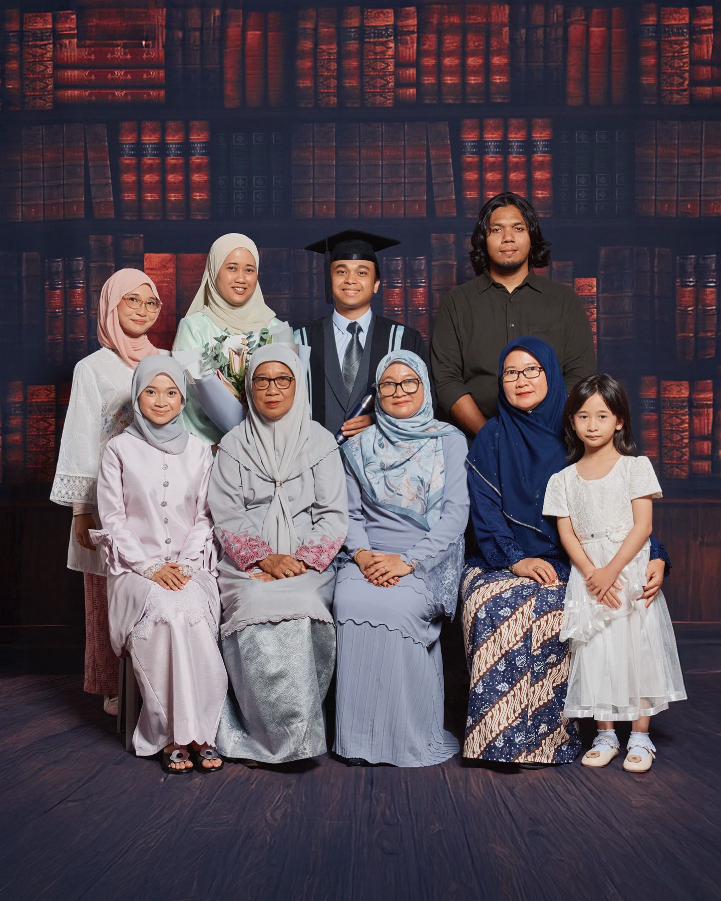 Muslim family portrait with graduation student in cap and gown posed together in front of bookshelf backdrop.