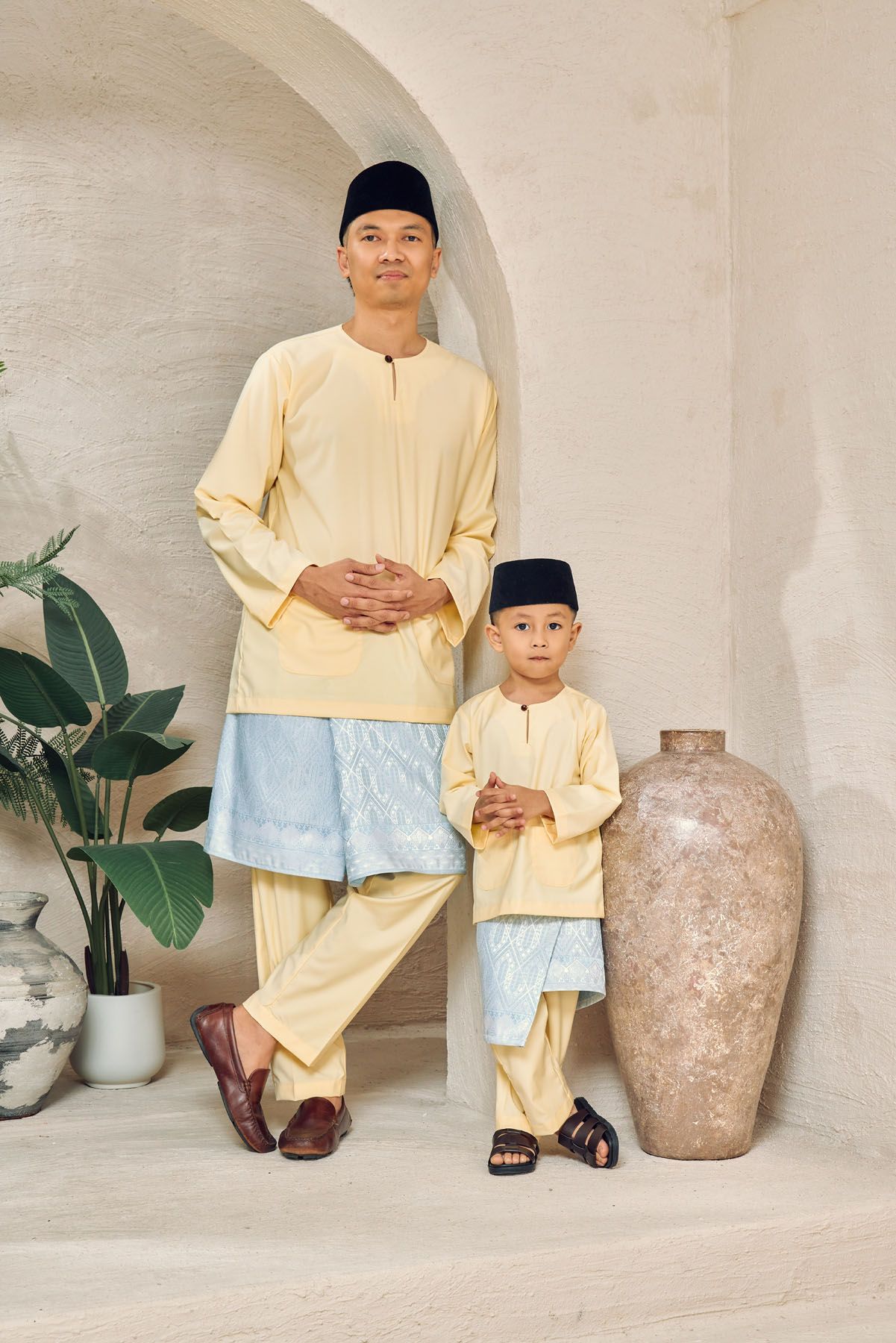 Malaysian father and son in matching yellow baju melayu with blue songket standing together.