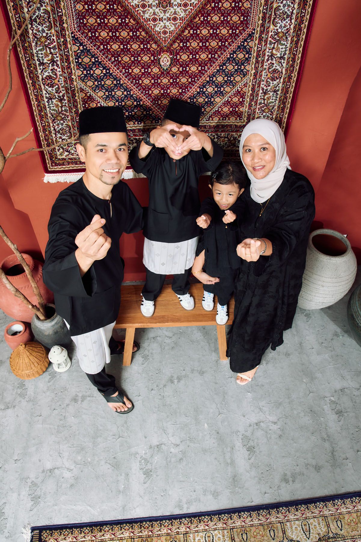Malaysian Muslim family in black traditional attire making finger heart gestures indoors.