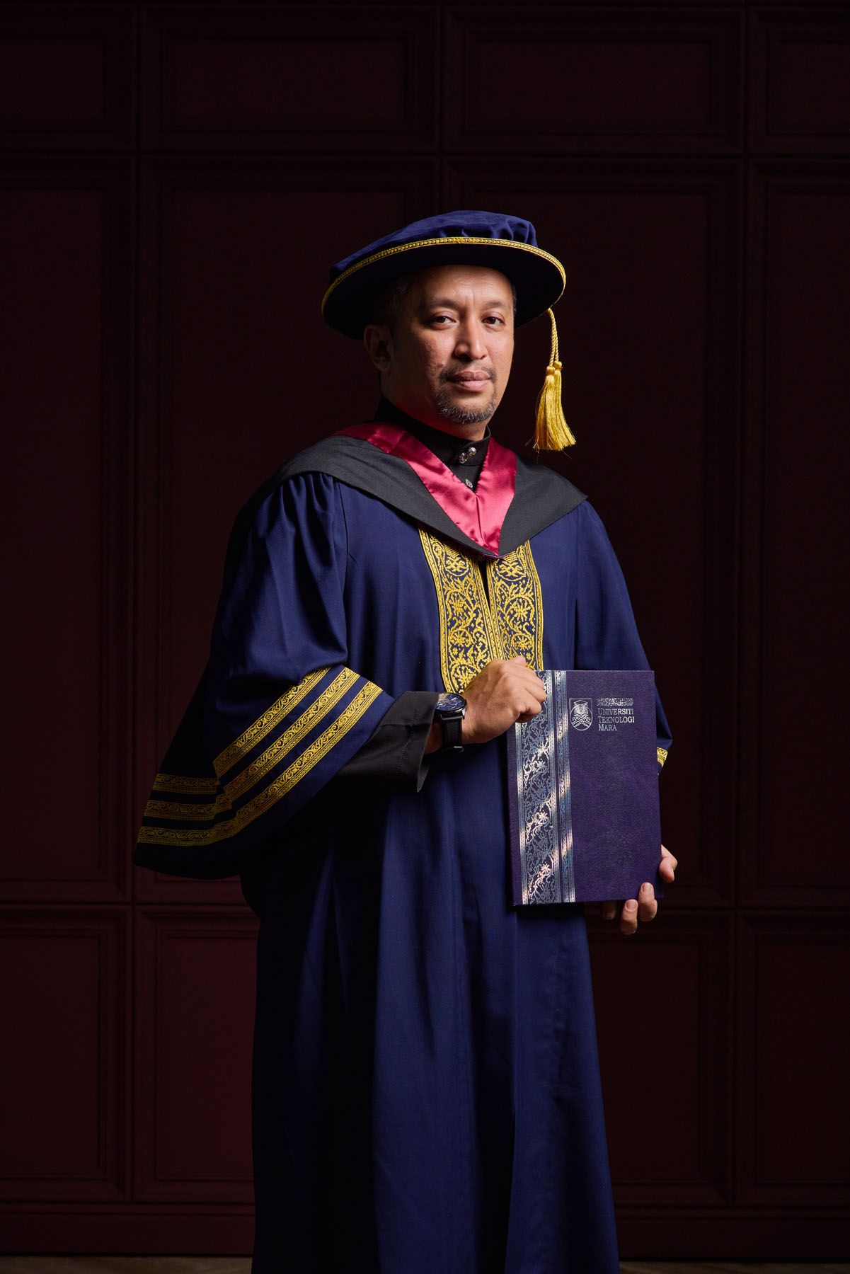 Distinguished academic in blue doctoral graduation robes with gold trim holds book before dark wood panel background.