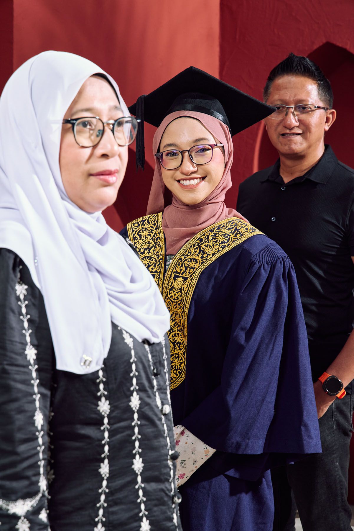 Muslim graduate in navy gown and mortarboard smiles with proud parents at graduation ceremony.