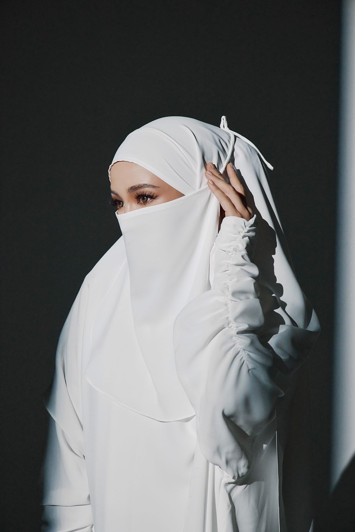 Muslim woman in white niqab and hijab covering face, posing against dark background in modest attire.
