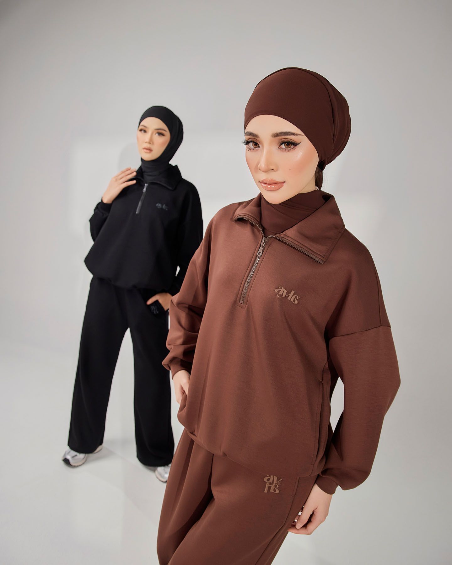 Two women modeling brown and black modest zip-up sweatsuit sets with hijabs in a studio setting.