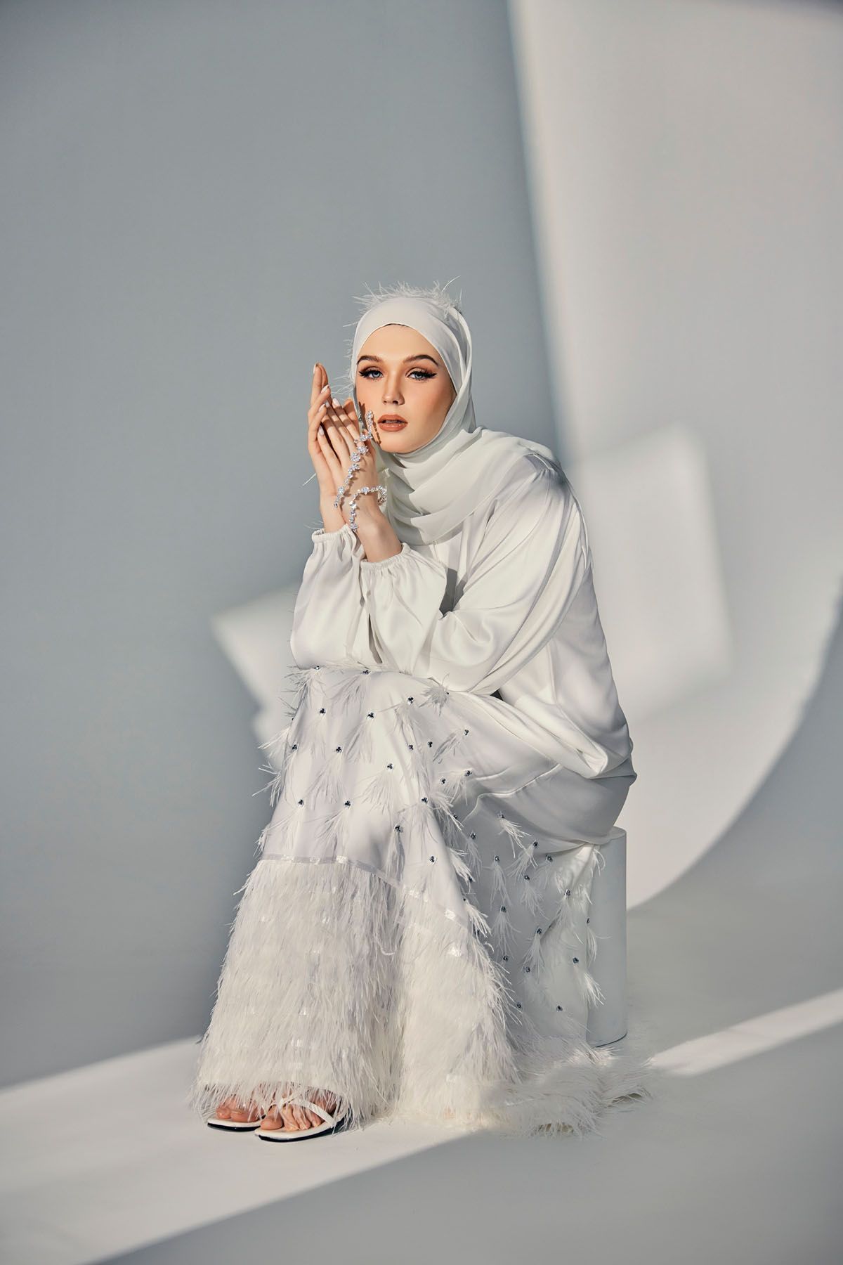 Woman in white feather-trimmed hijab abaya seated gracefully with hands clasped in a white studio.