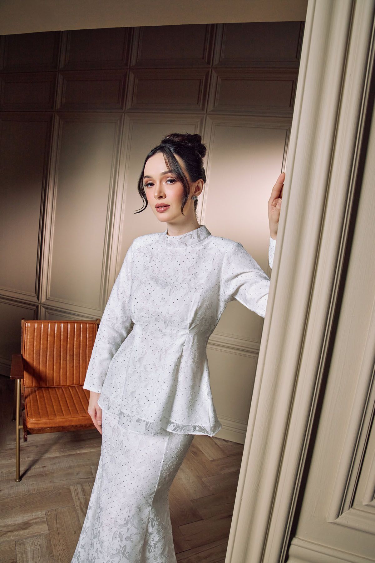 Woman in white lace peplum baju kurung stands in classic interior.