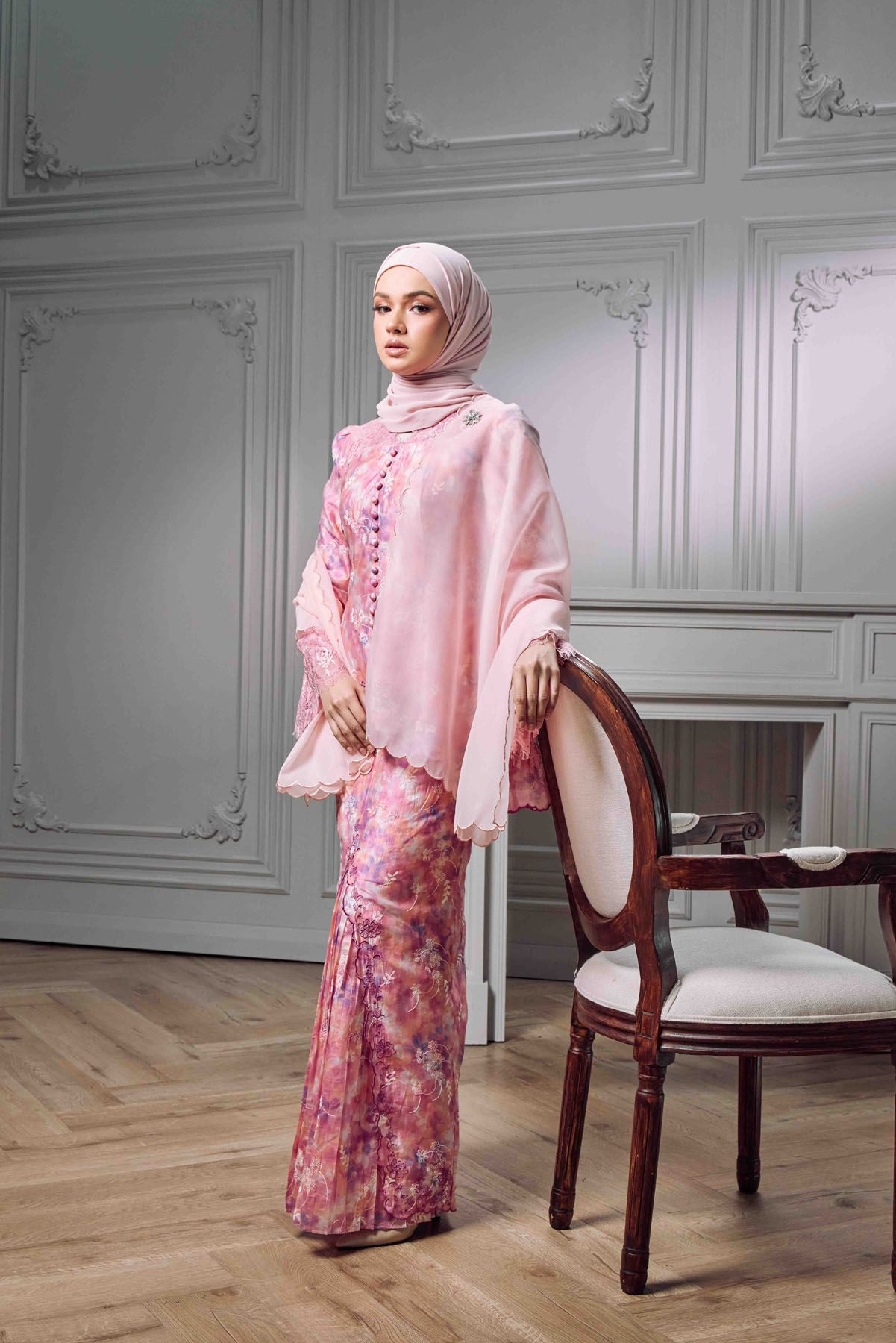 Muslim woman in pink baju kurung and hijab standing gracefully beside an ornate chair in a white room.