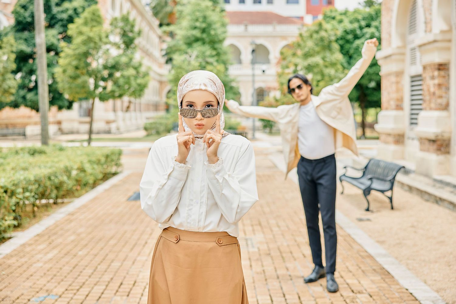 Two women in hijabs pose playfully on a brick pathway in a sunny outdoor urban environment.