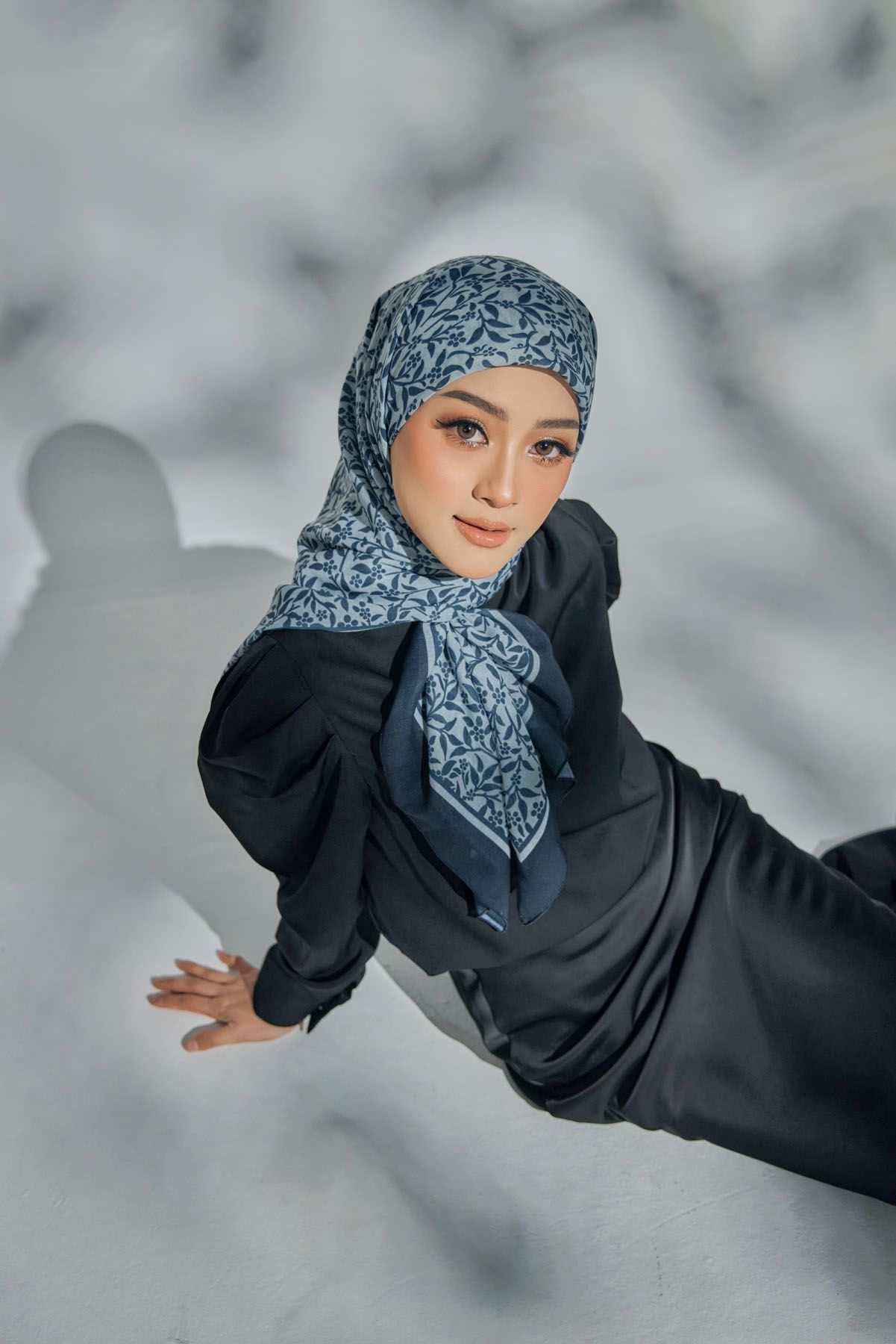 Aerial view of woman in black outfit and blue patterned hijab posing on white background.