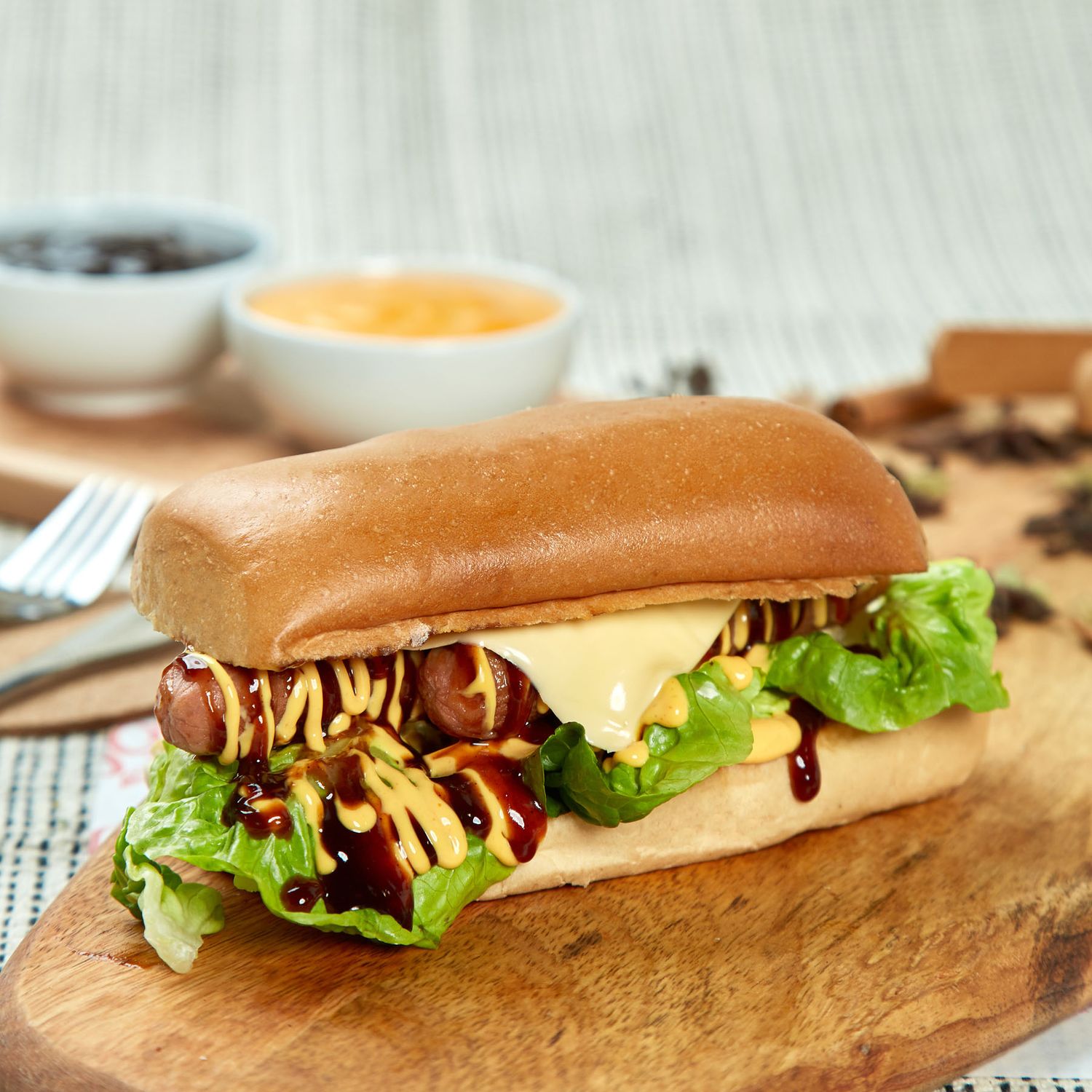 Hot dog sandwich with lettuce, mustard, BBQ sauce in brioche bun on wooden board with dipping sauces.