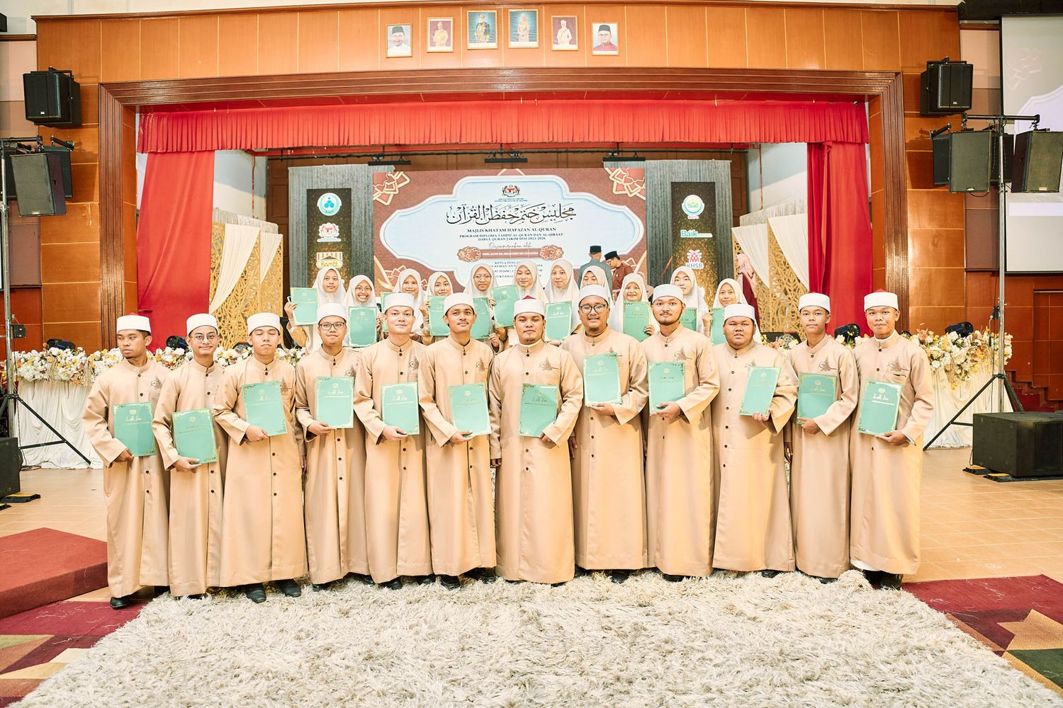 Group of male Islamic students in traditional white attire holding certificates at a formal Quran graduation ceremony.