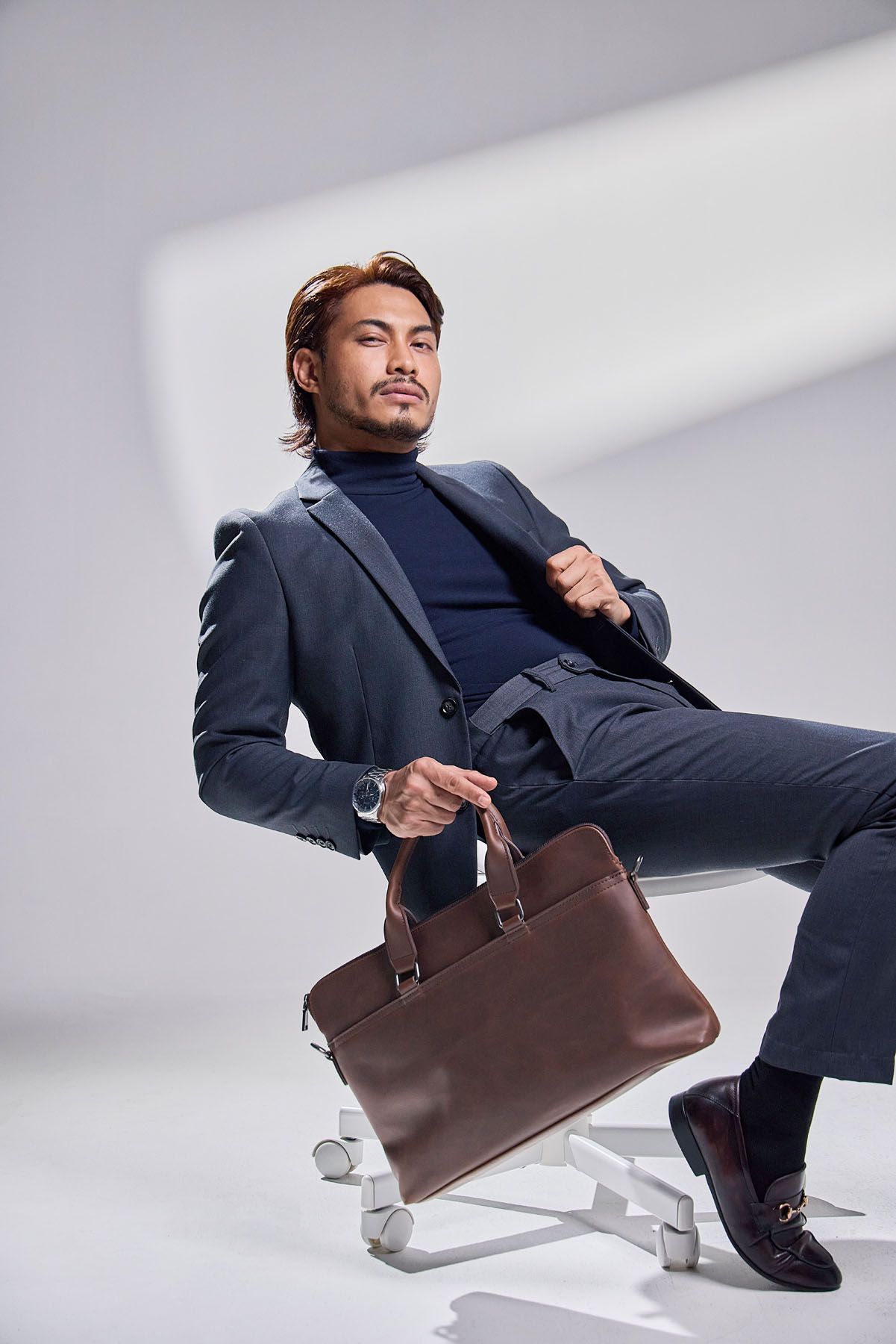 Stylish Asian man in navy suit holding brown leather briefcase while seated on chair.