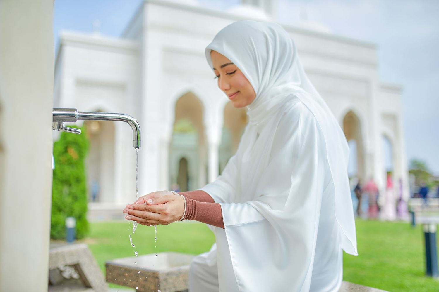 Muslim woman in white hijab performing wudu ablution washing hands at outdoor mosque fountain.