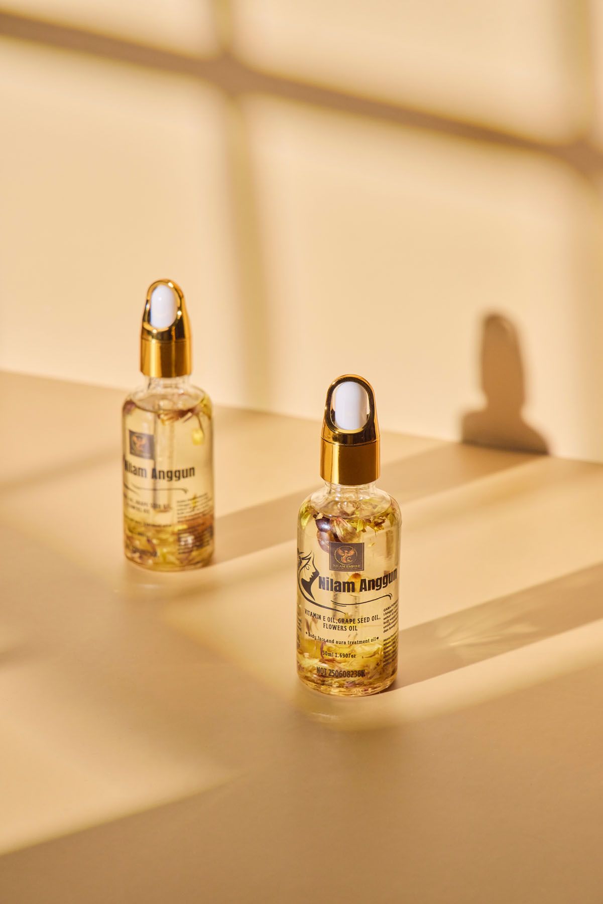 Two Hilam Anggun vitamin E facial serum dropper bottles with gold caps on a warm beige background.