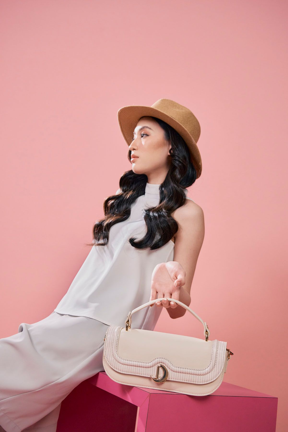 Asian woman in white sleeveless top and hat holding a cream structured handbag on pink background.