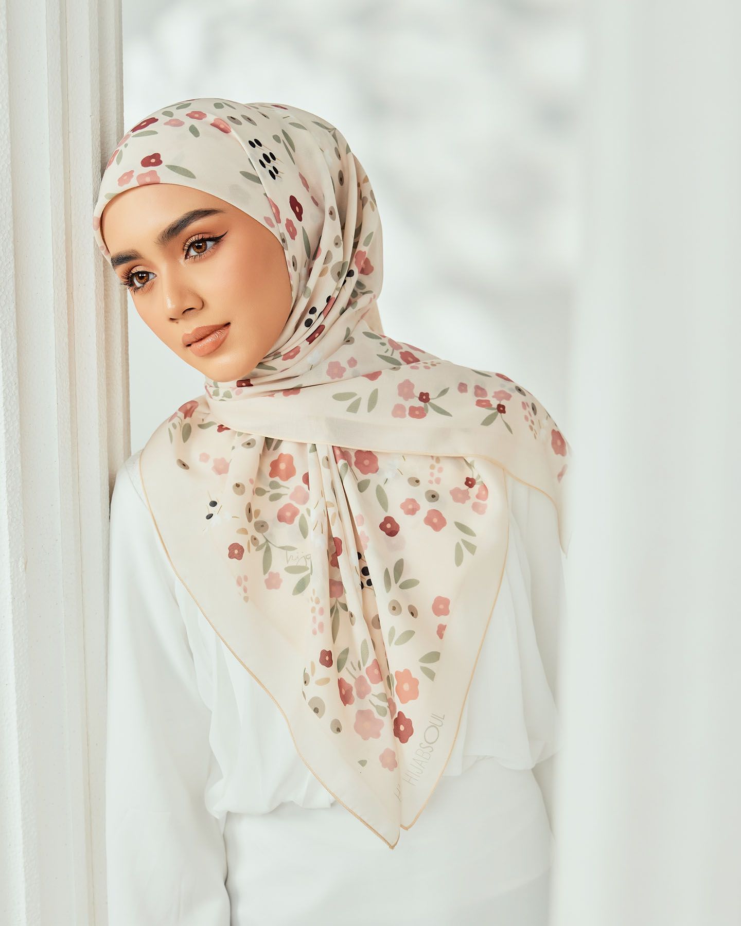 Woman wearing a floral printed square hijab in cream with pink roses near a bright window.