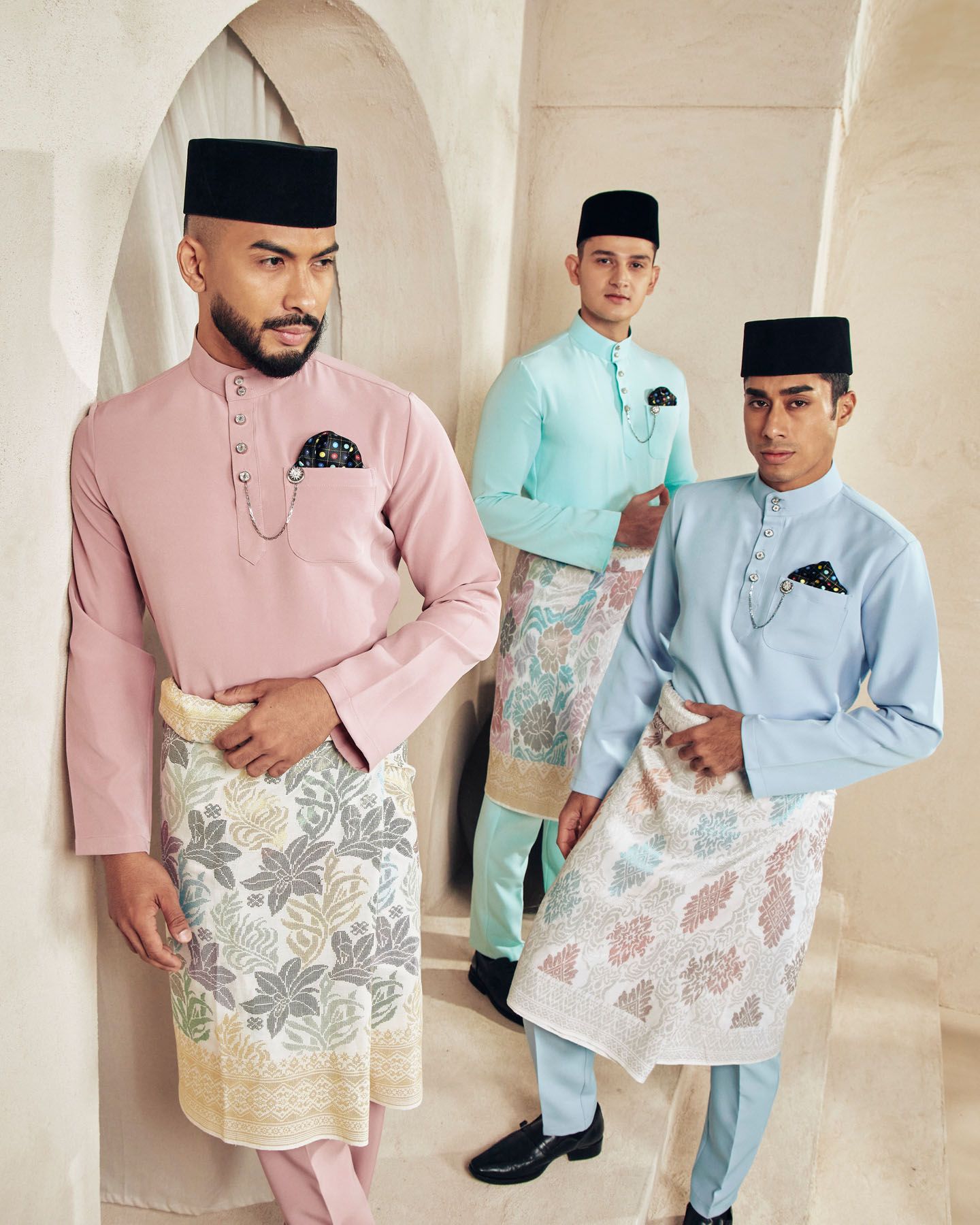Three men in pastel baju melayu with black songkok and floral sampin pose together.