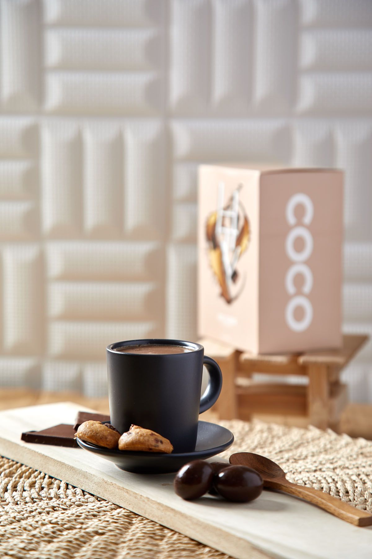 Dark mug of hot chocolate with cookies and chocolate pieces beside a Coco brand box on a woven mat.