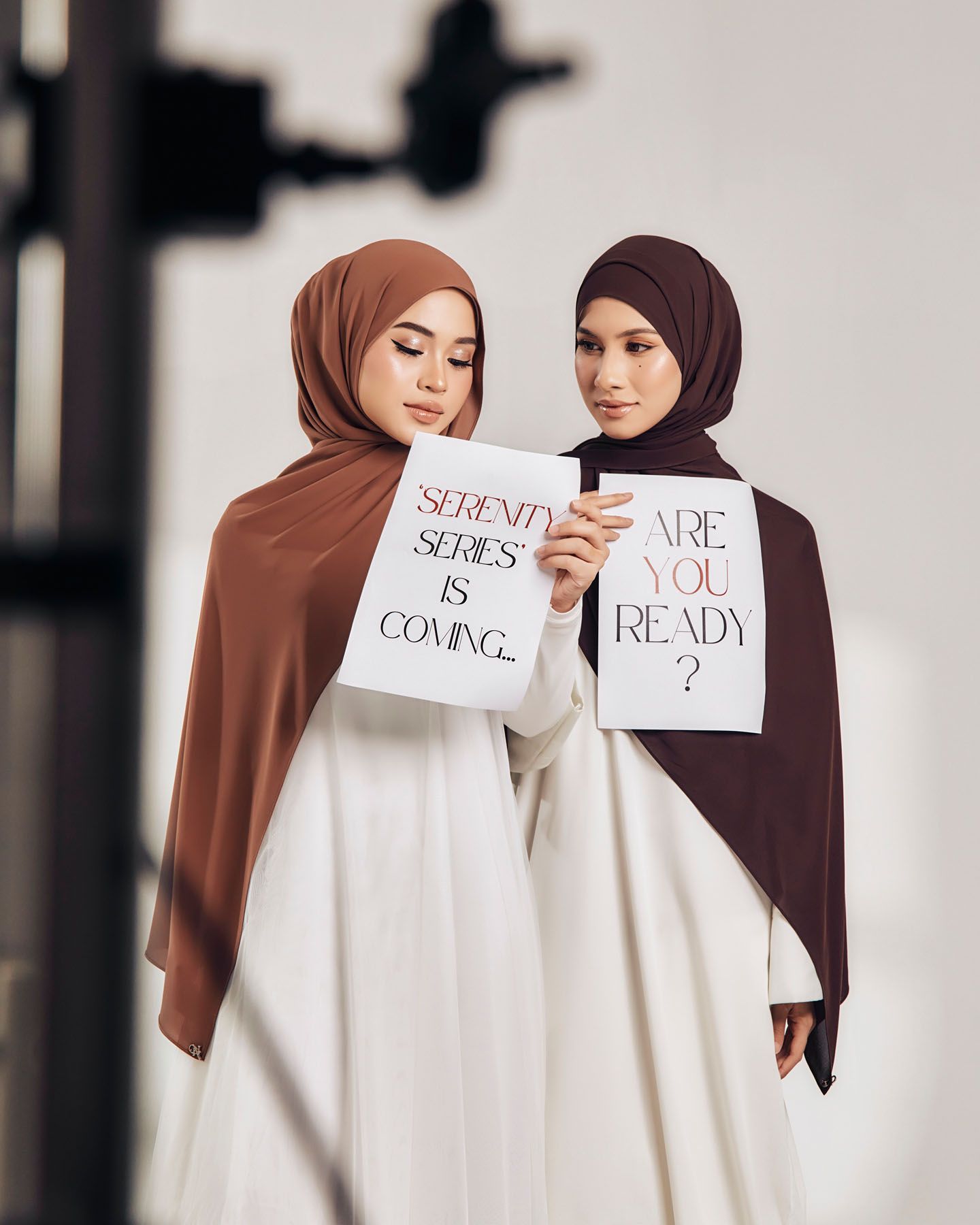 Two women in hijabs hold signs reading Serenity Series Is Coming and Are You Ready during a fashion campaign shoot.