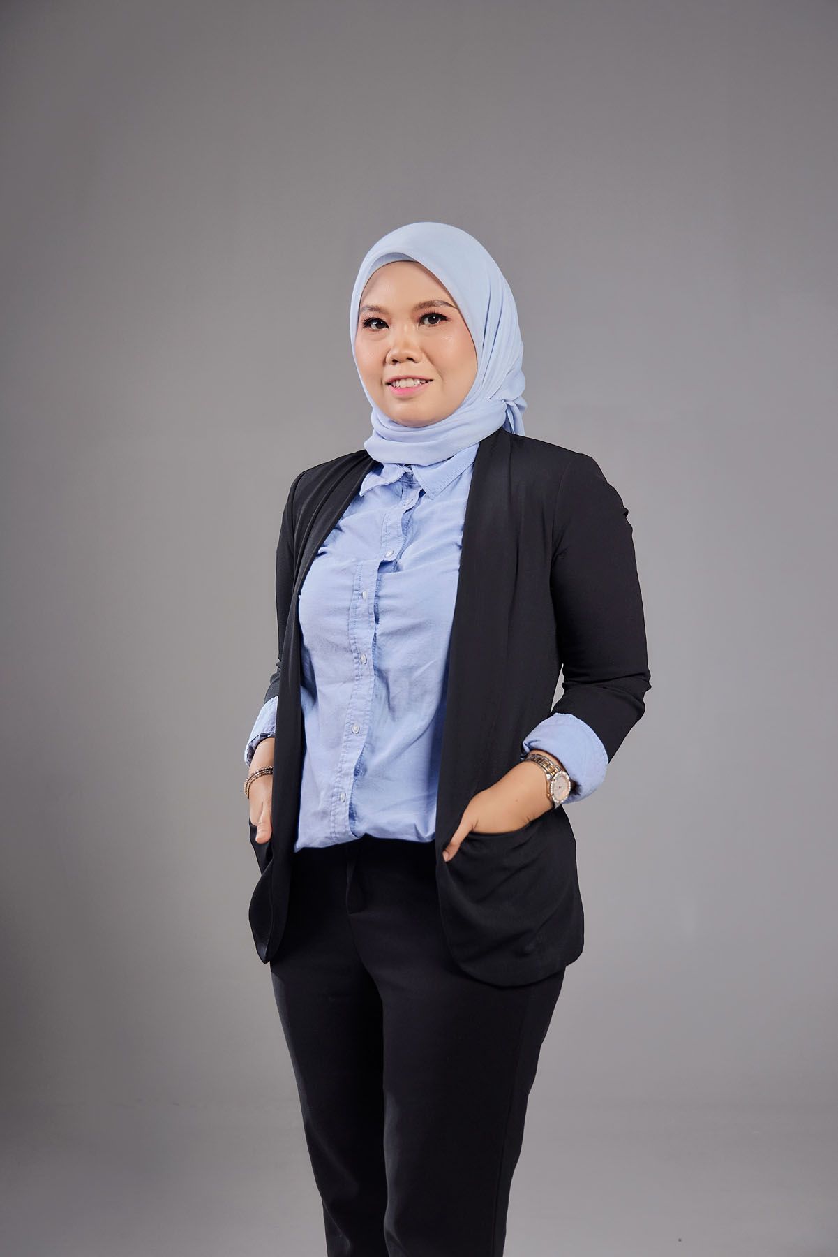 Confident Muslim businesswoman in light blue hijab and black blazer standing with hands in pockets.