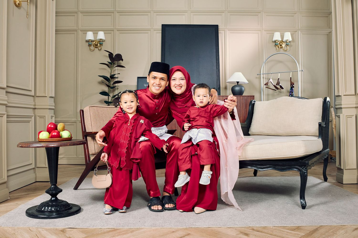 Young Muslim family in coordinating red Baju Raya traditional outfits posing in stylish interior.