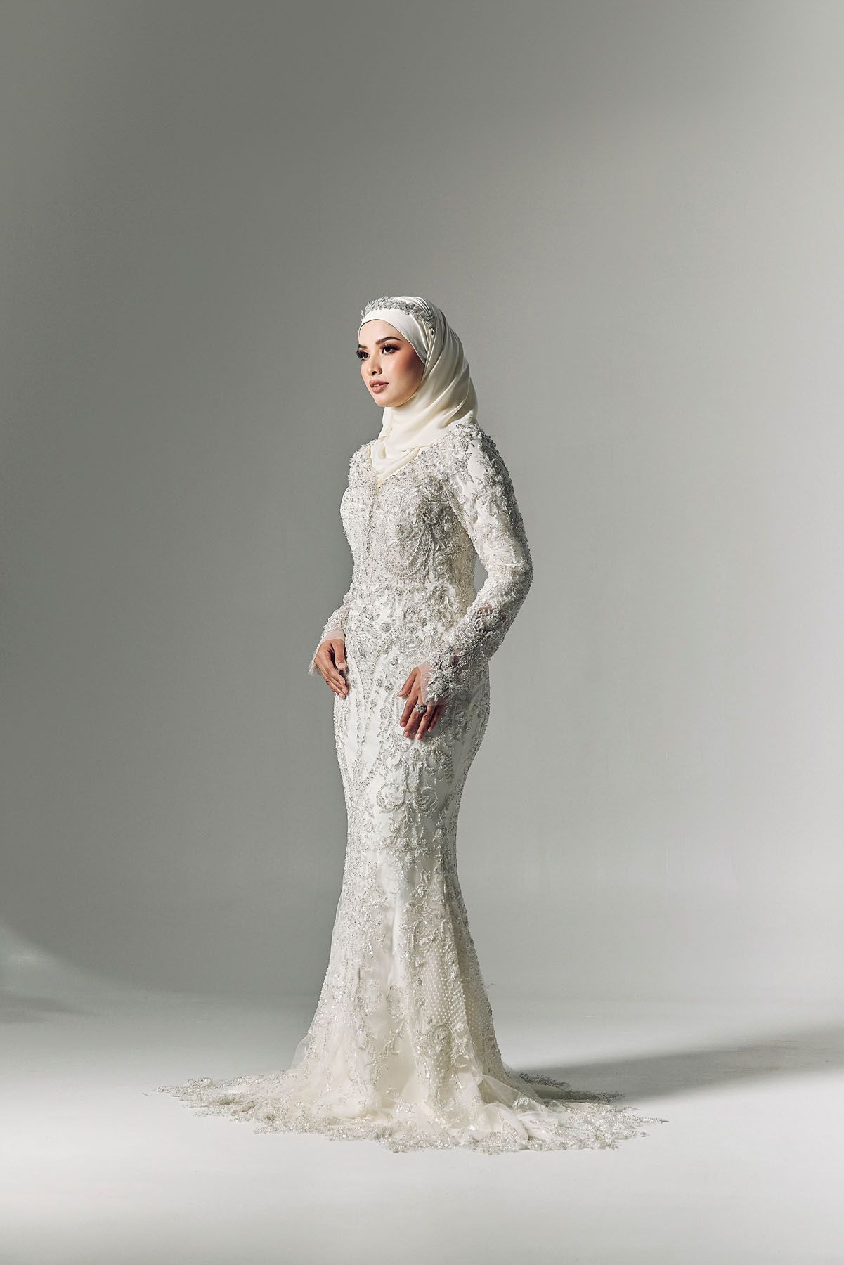 Muslim bride in a fitted white lace wedding gown with matching hijab posing elegantly on a grey background.