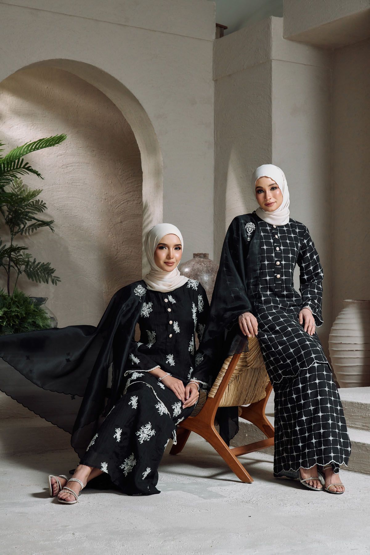 Two Muslim women in matching black embroidered baju kurung sets with white floral patterns and white hijabs.