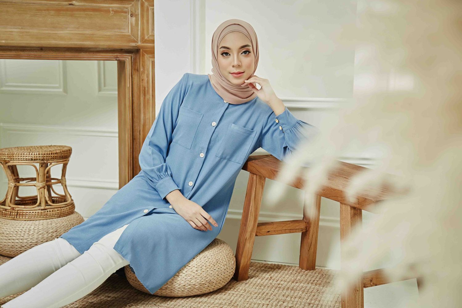 A woman in a blue modest dress and nude hijab sits relaxed on a rattan cushion in a cozy wooden interior.