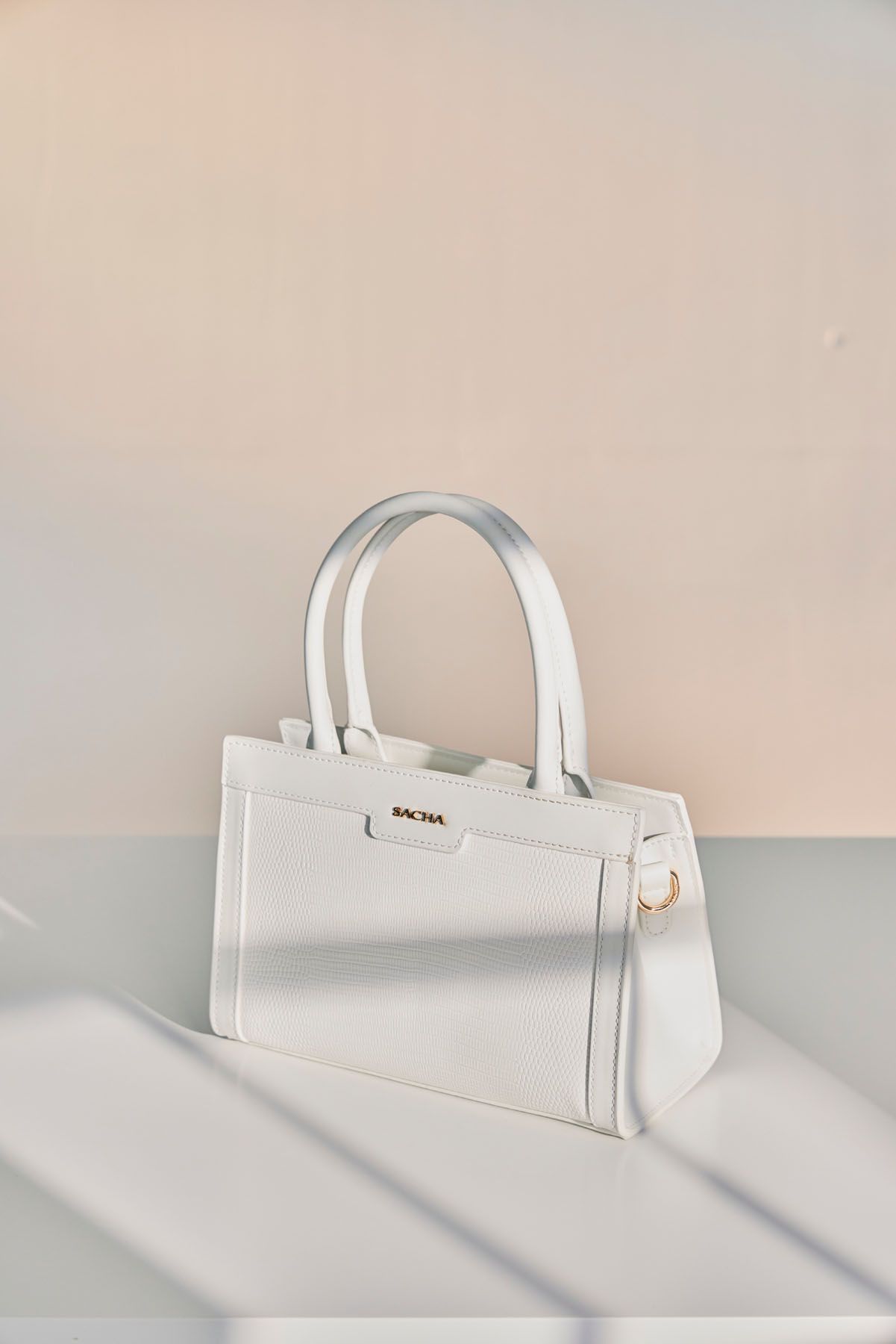 Elegant white structured leather Sacma handbag with gold hardware displayed on a white shelf in soft light.