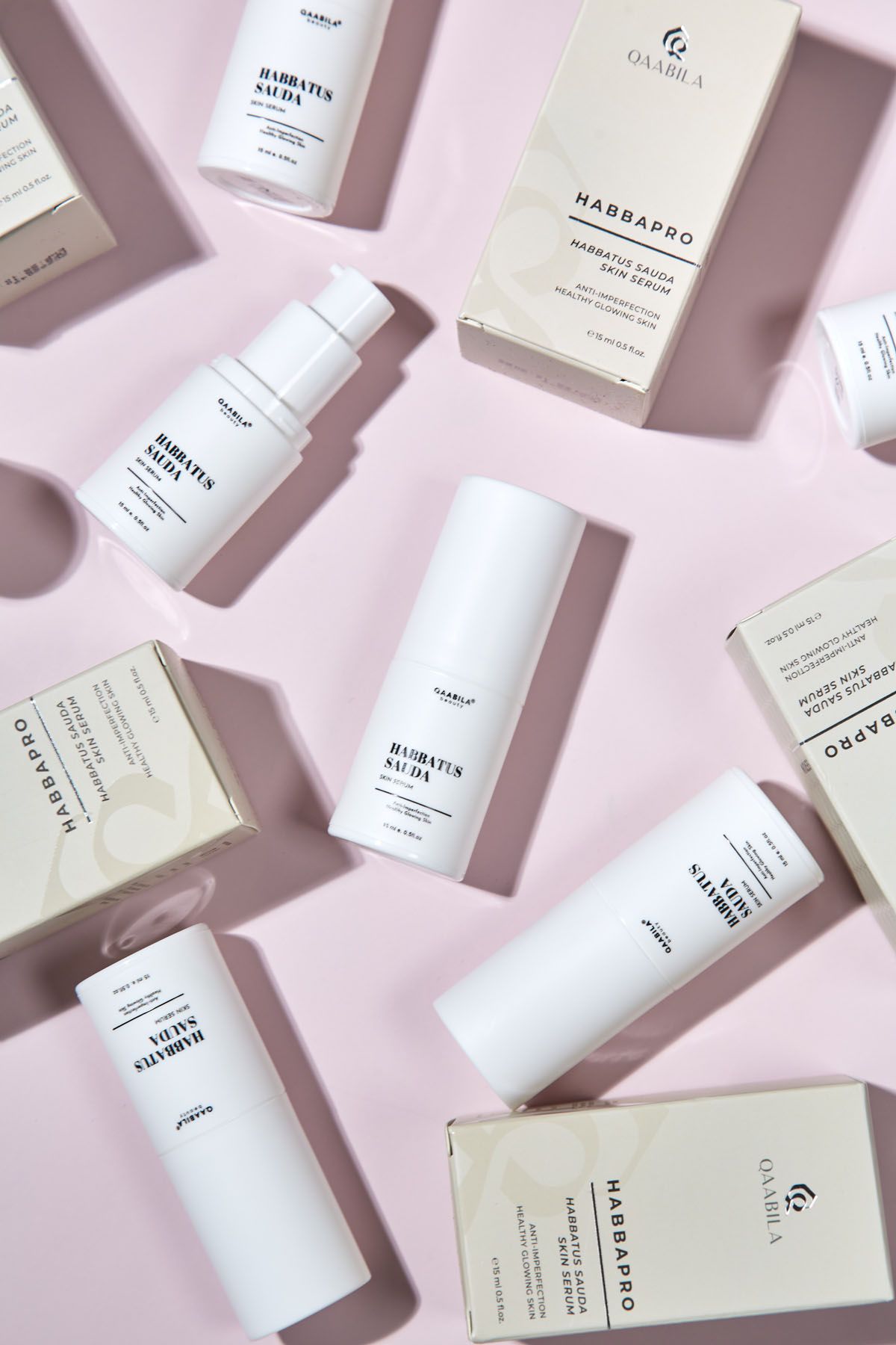Flat lay of Qaabila Habbapro and Habbatus Sauda skincare bottles and boxes scattered on pink background.