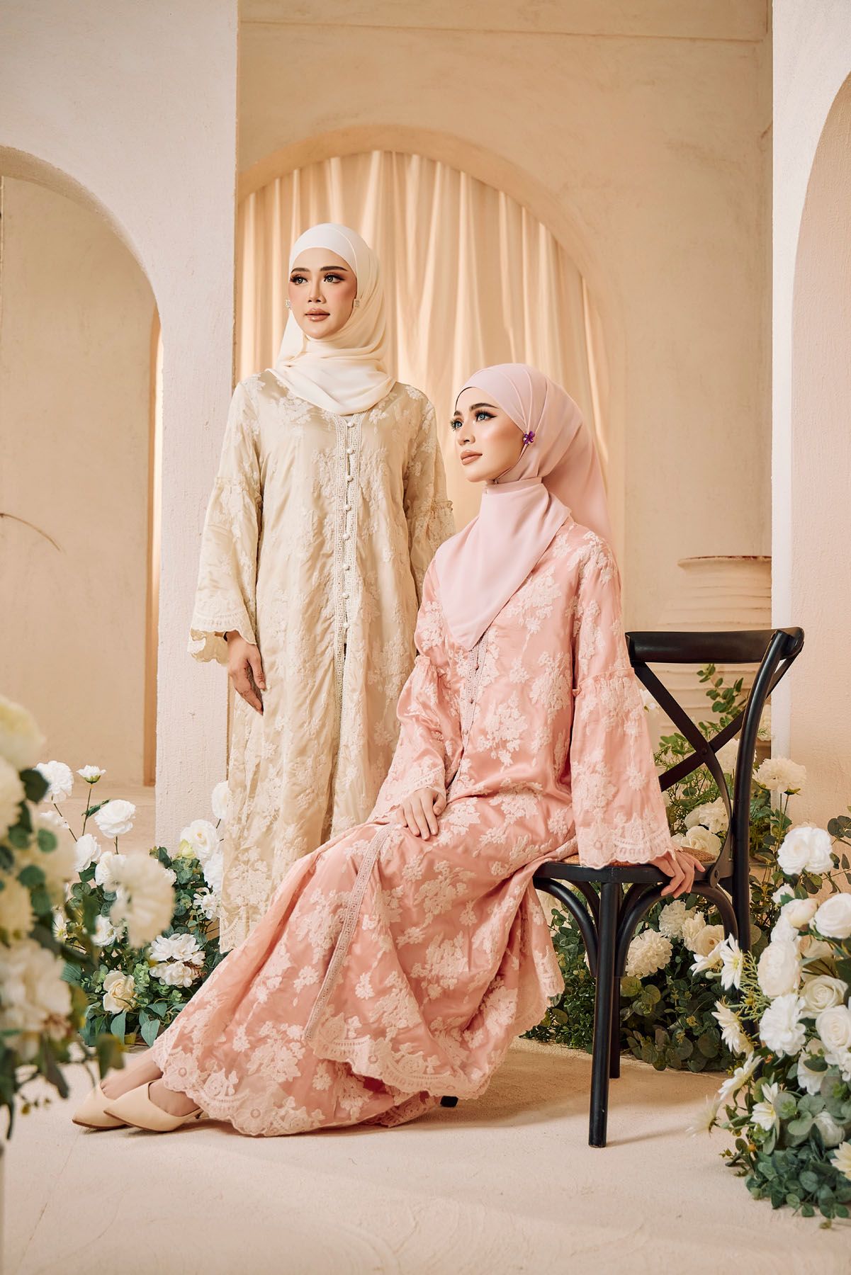 Two women in elegant floral embroidered abayas in gold and pink pose among white flowers.