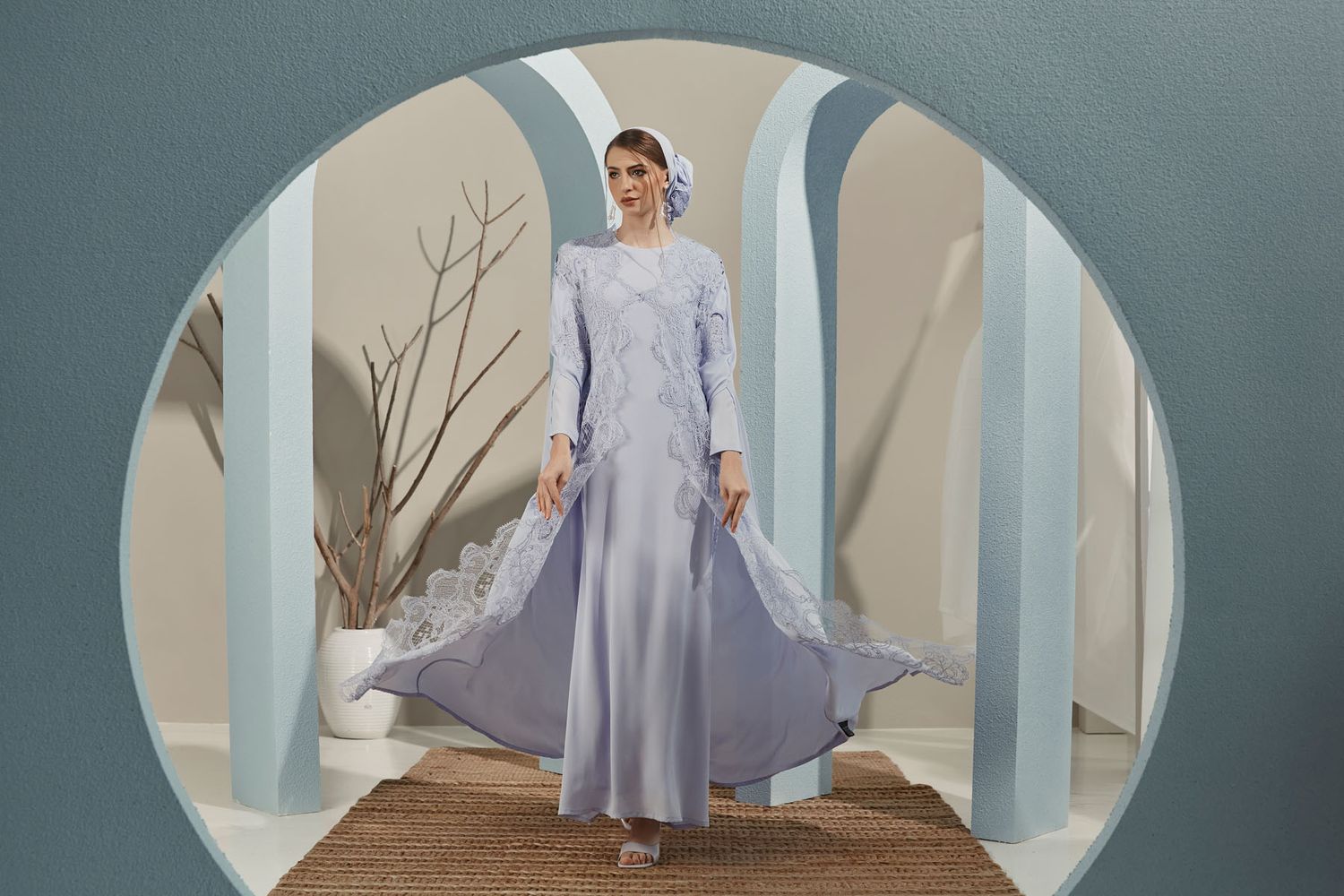Woman in flowing light blue lace abaya and floral hijab posing inside circular arch doorway.