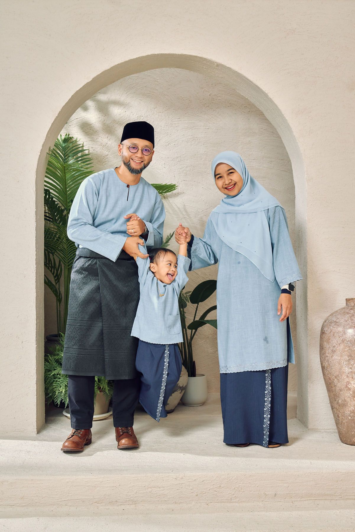 Muslim family of three in matching light blue traditional Malay baju kurung and baju melayu outfits with young child.