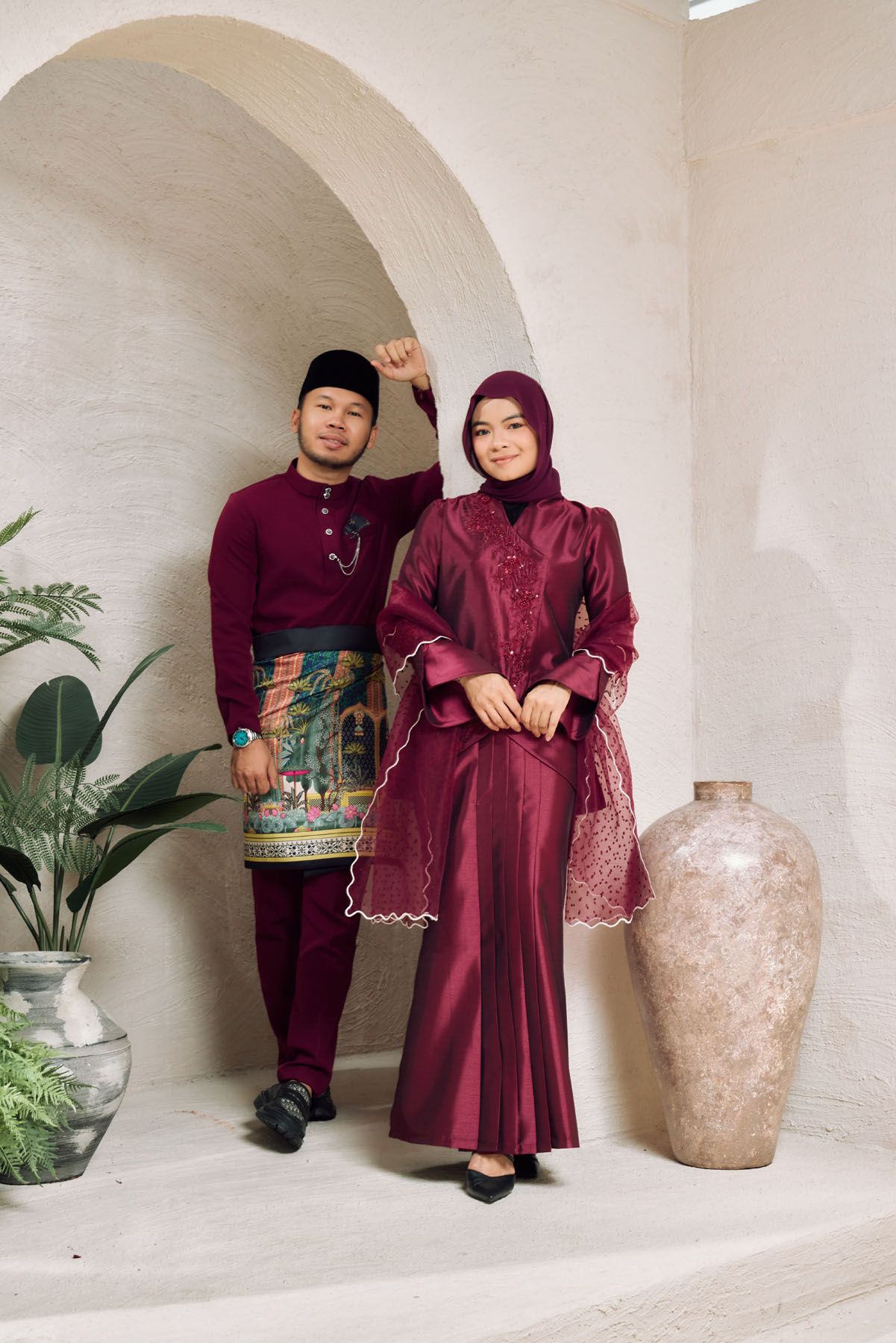 A Malaysian couple in matching maroon traditional Raya outfits posing beside a large ceramic vase under an arch.