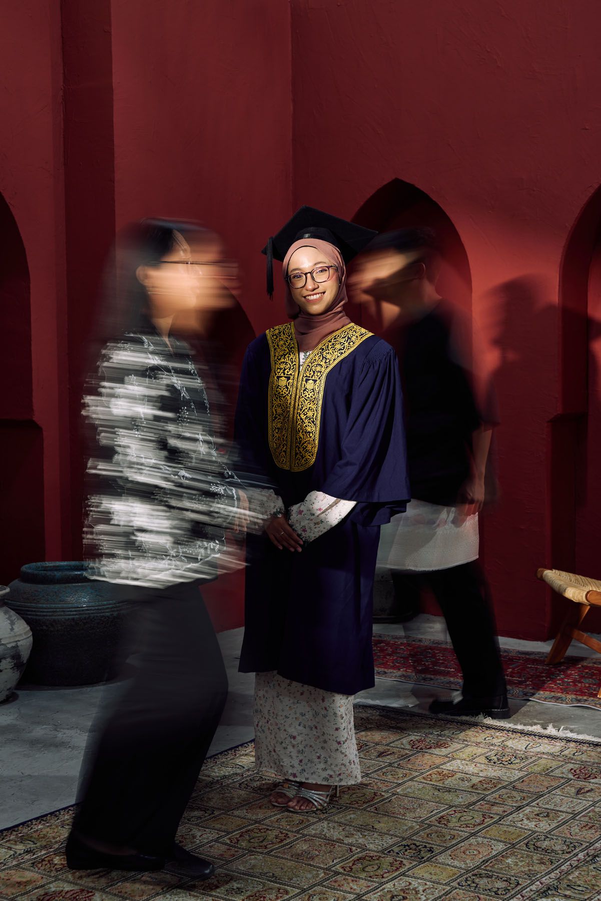Muslim graduate in navy gown stands still as blurred figures move around her.