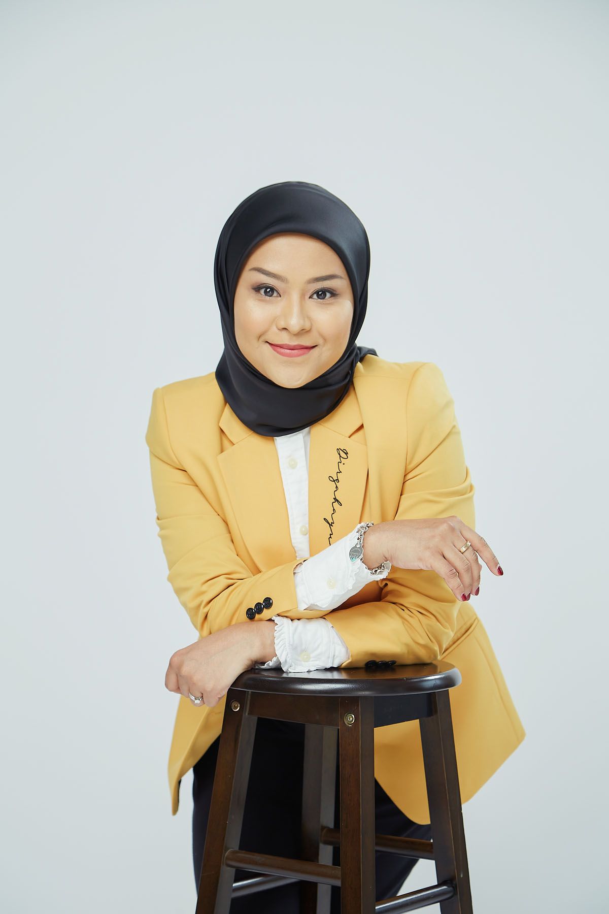 Confident Muslim businesswoman in yellow blazer and black hijab leaning on wooden stool smiling.