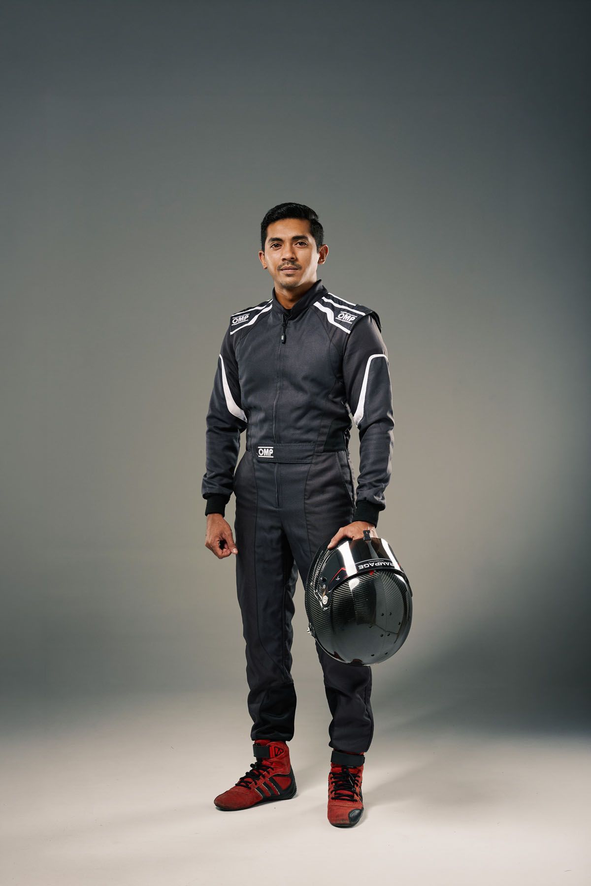 Male racing driver in black OMP suit holding helmet, wearing red racing shoes, posing in studio.