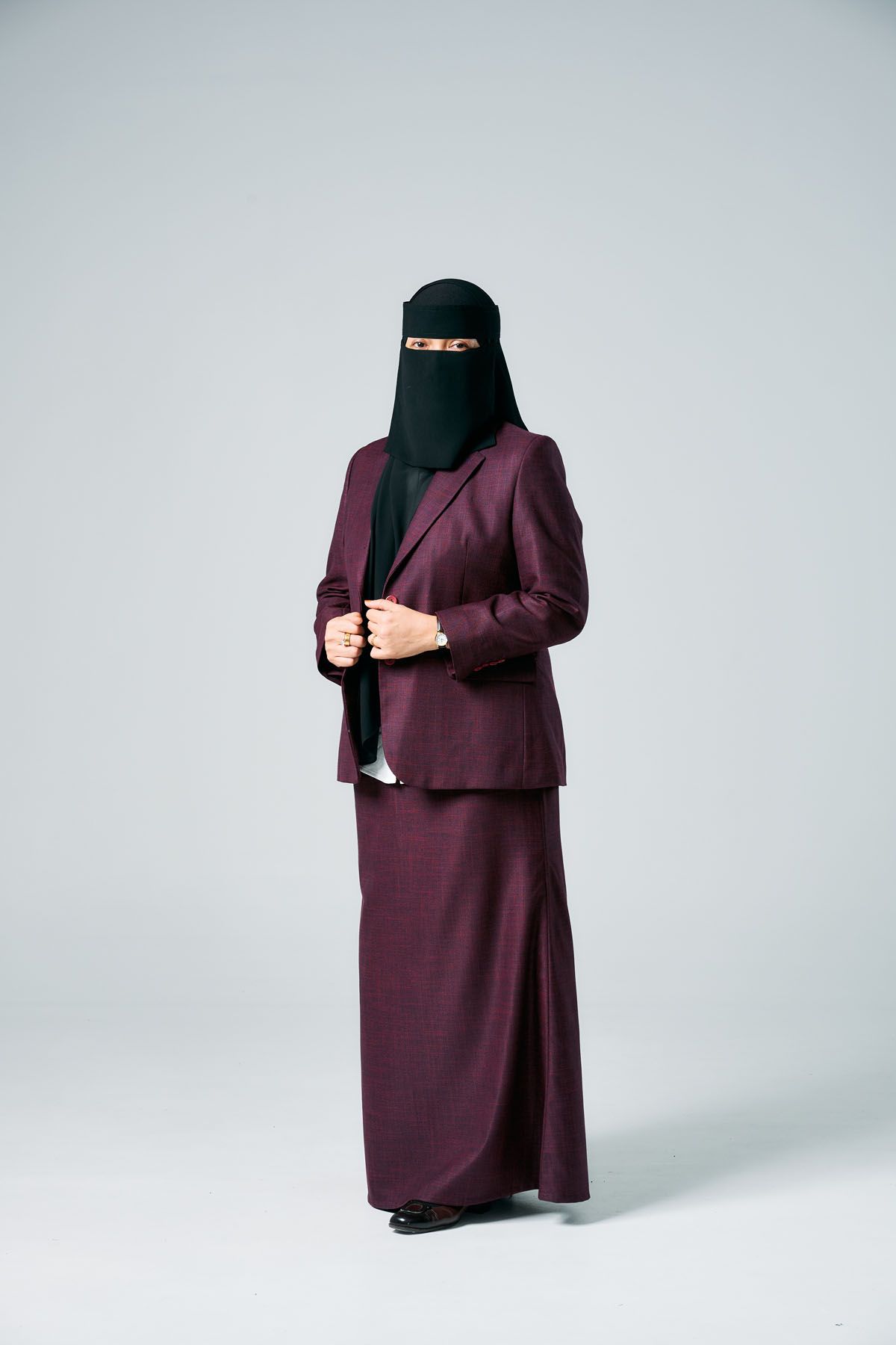Muslim woman in burgundy suit and black niqab standing against gray background.