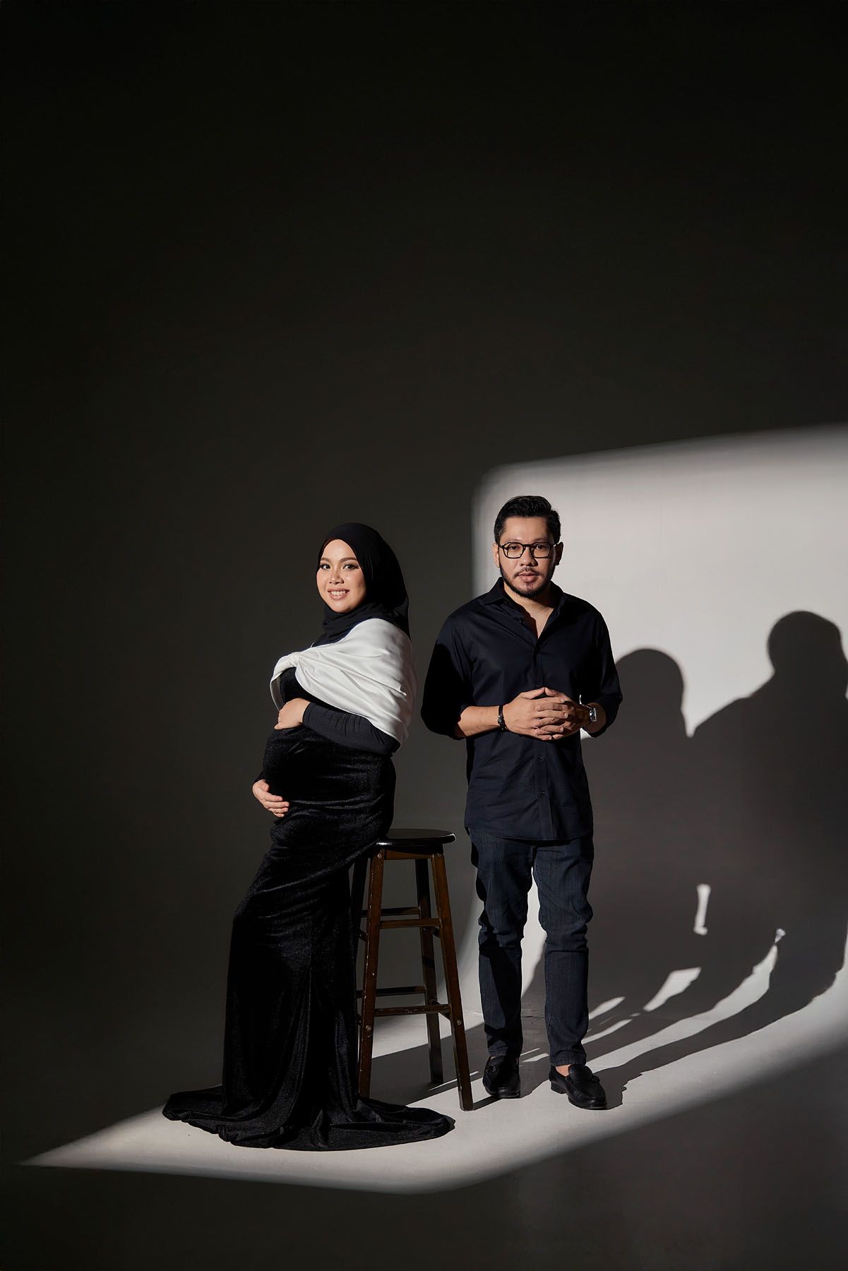 Pregnant Muslim woman in black hijab and gown poses with husband in dark studio with dramatic lighting.
